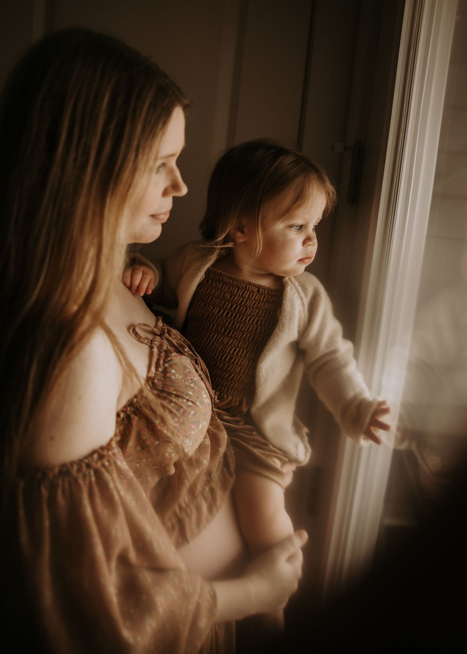 Two figures stand by a sunlit window in a warm, sepia-toned scene wearing light colored clothing.
