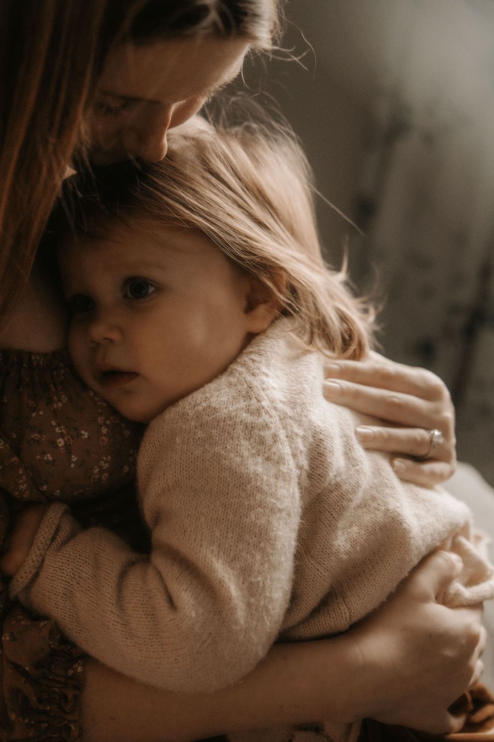 Intimate moody portrait of parent and child embracing in soft natural light wearing neutral tones.