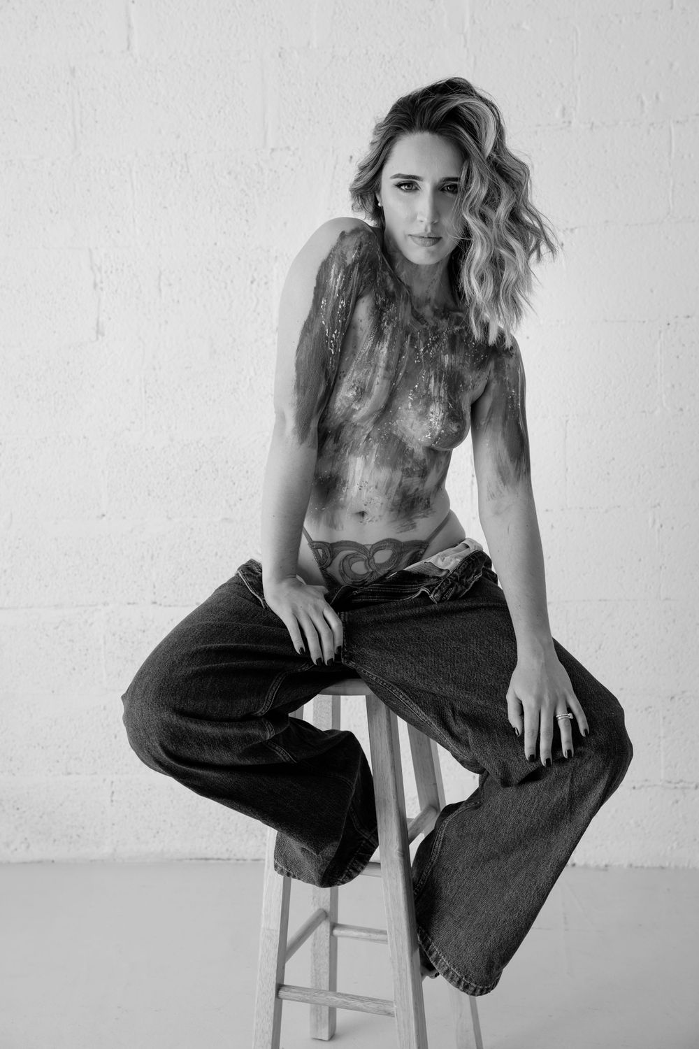 Artistic black and white portrait of person in jeans sitting on stool against white wall.