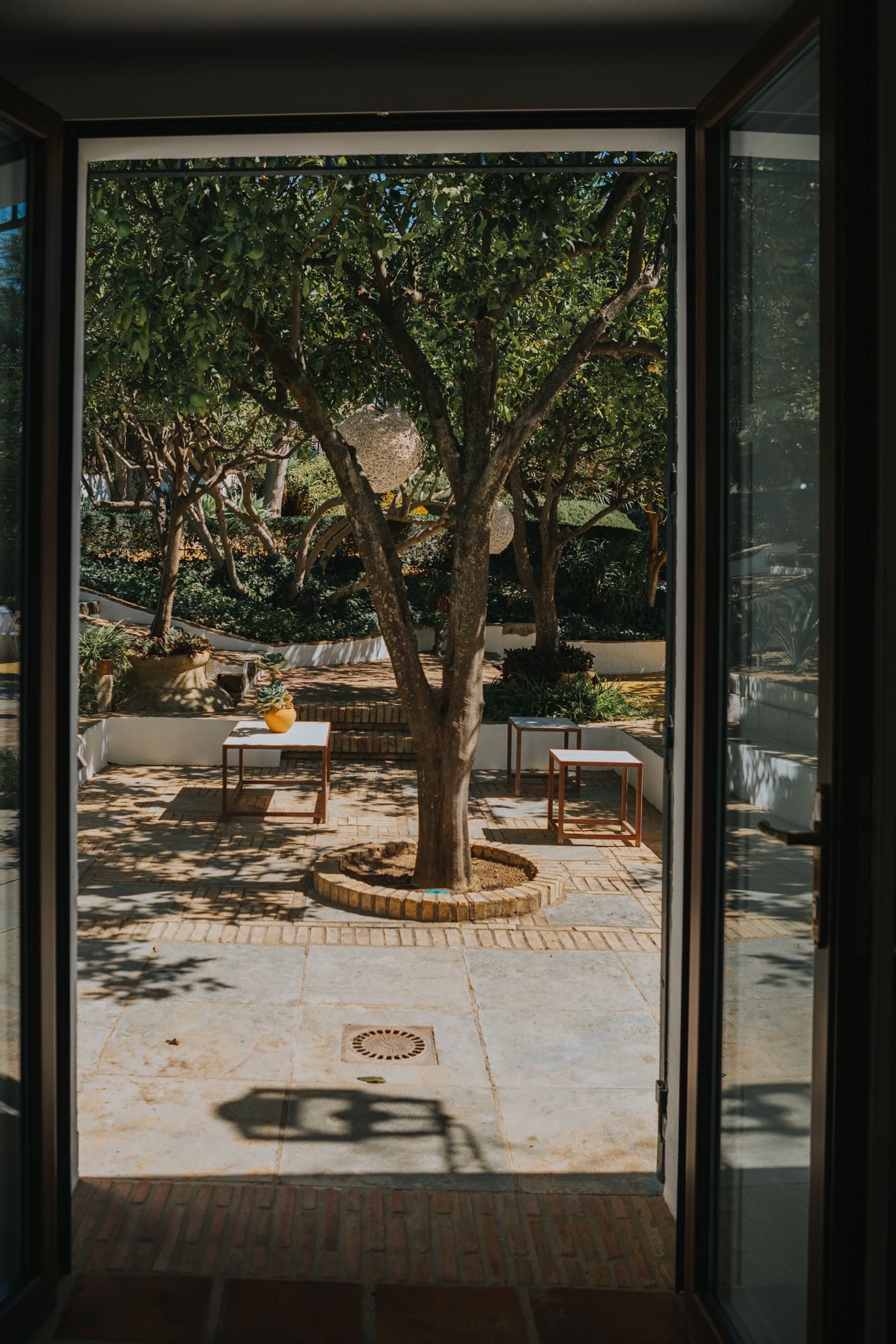 View through doorway of sunny courtyard with tree, brick paving, and outdoor seating area creating dappled shadows.