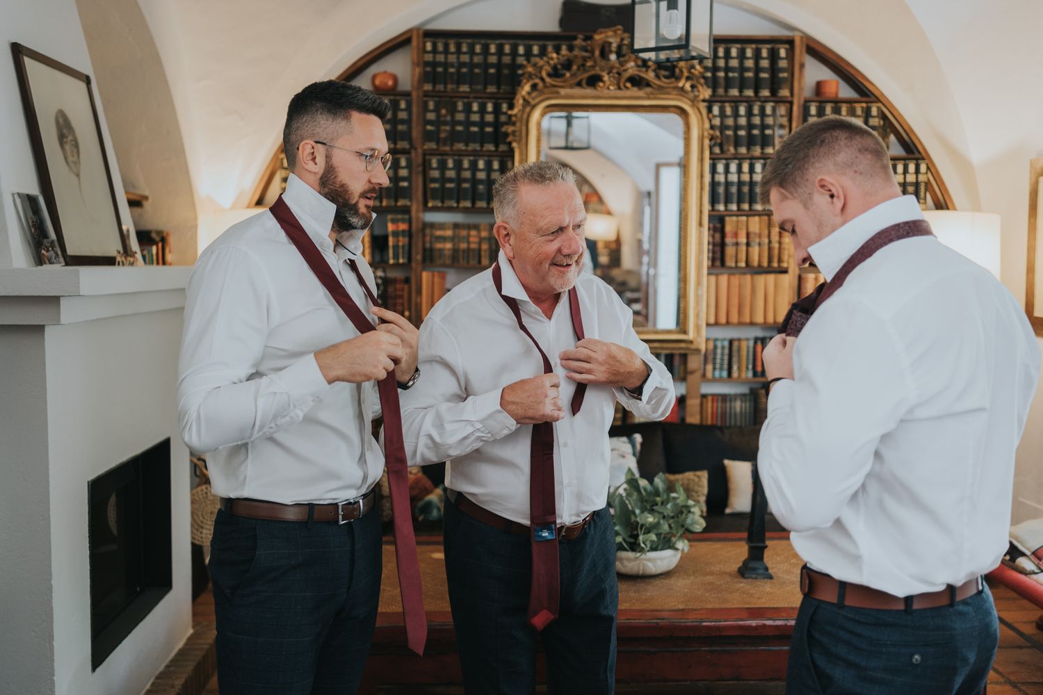 Three people in white shirts and suspenders help each other get dressed in a vintage-style room with ornate wallpaper.