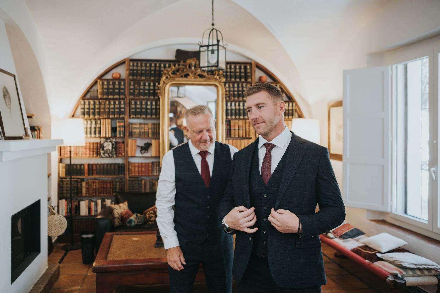 Two well-dressed men converse in an elegant library room with arched ceilings and wood bookshelves.