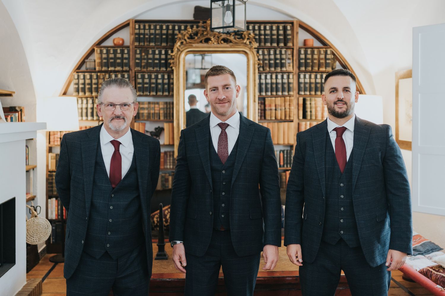 Three groomsmen wearing matching dark suits and red ties pose in an elegant room with arched bookshelves.