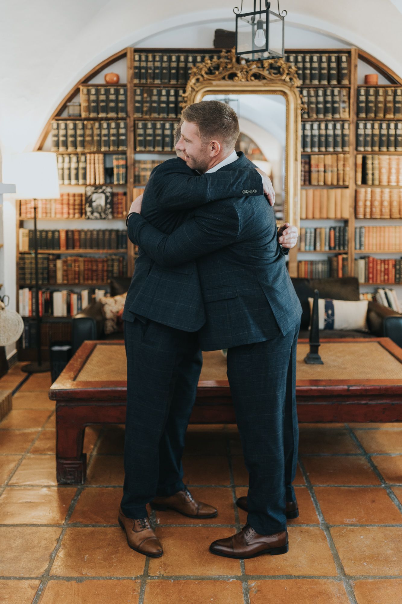 Two people in navy suits share an emotional embrace in a library setting with antique bookshelves.
