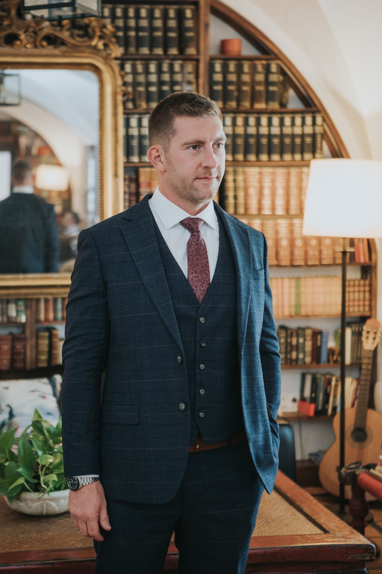 Elegant groom wearing a tailored navy three-piece suit and burgundy tie in a study with vintage decor.