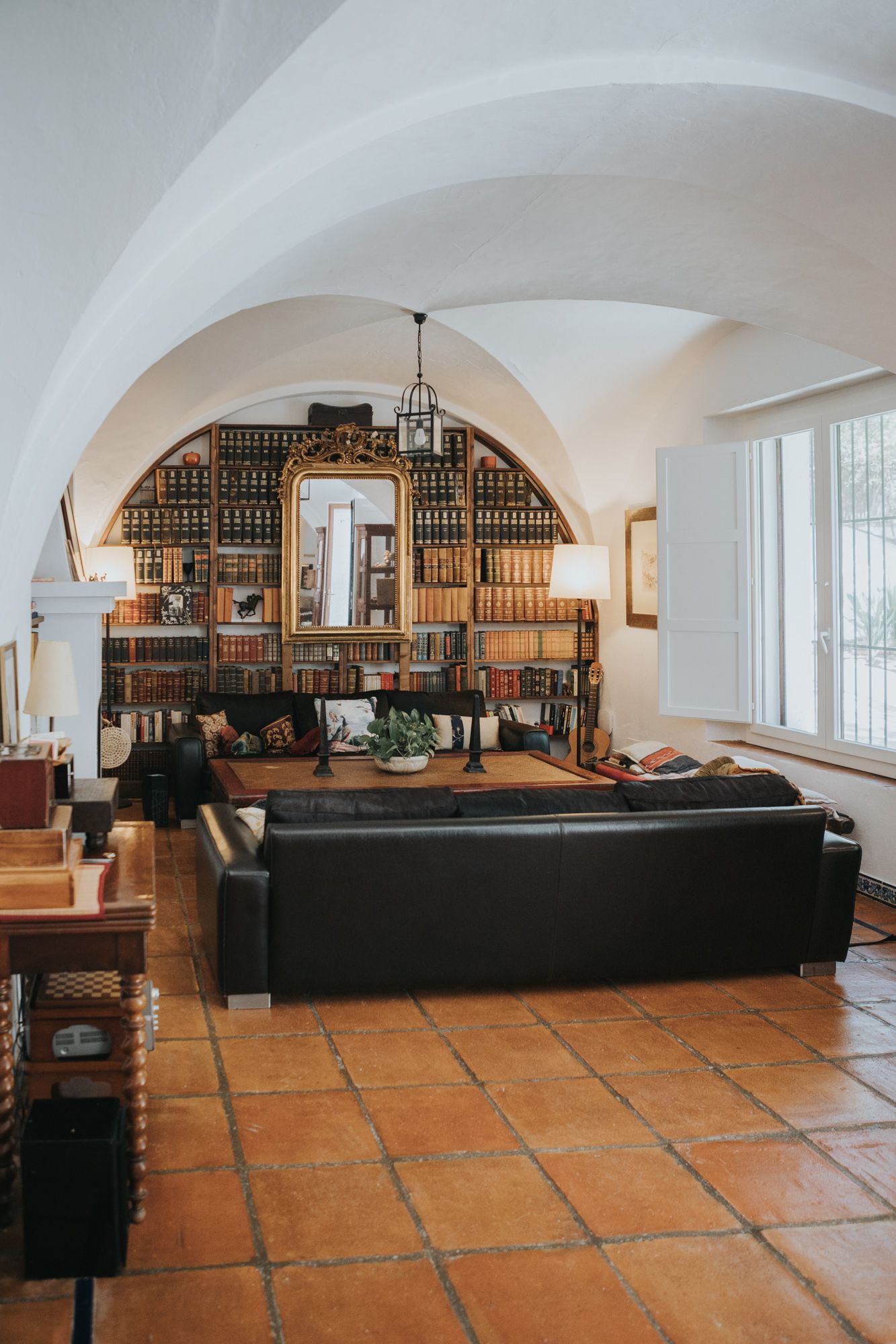 Elegant library room with arched ceiling, terra cotta floors, black leather sofa, and wooden bookshelves filled with books.