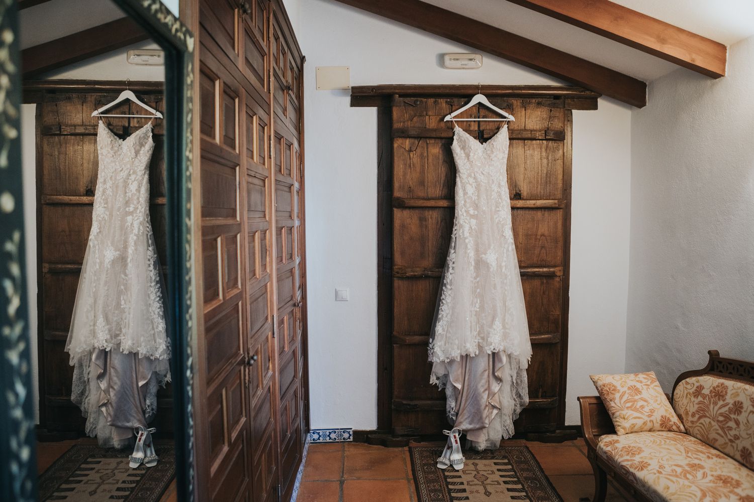 Elegant white lace wedding dress hangs on wooden doors in a rustic interior setting.