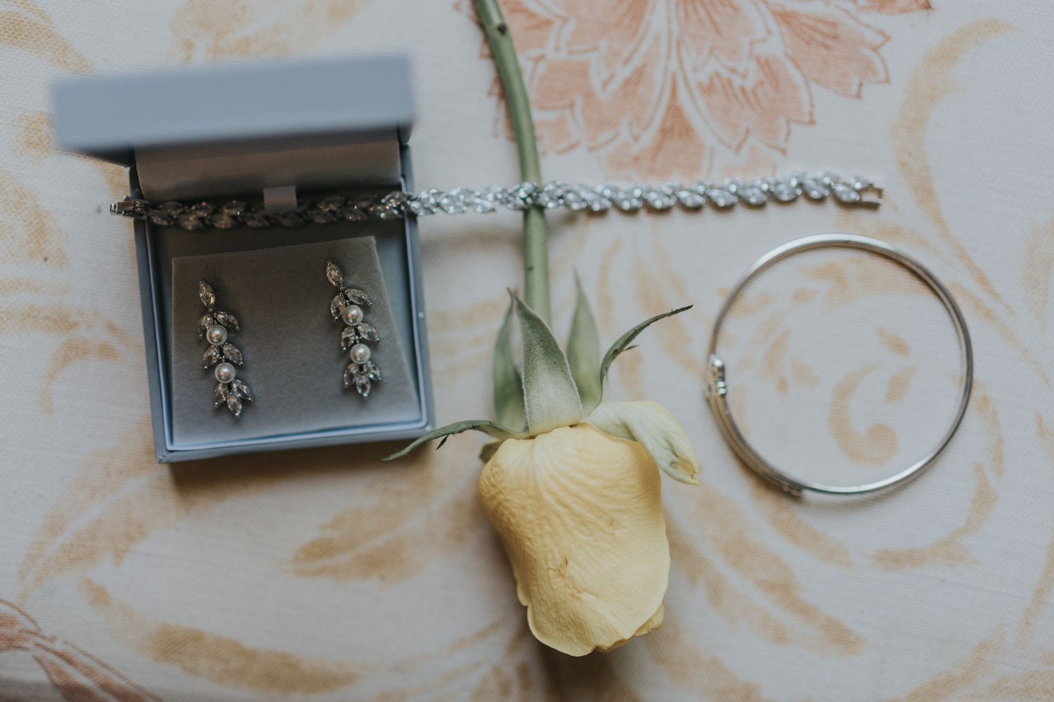 Wedding accessories including jewelry box, earrings, bracelet and a pale yellow rose on patterned fabric.