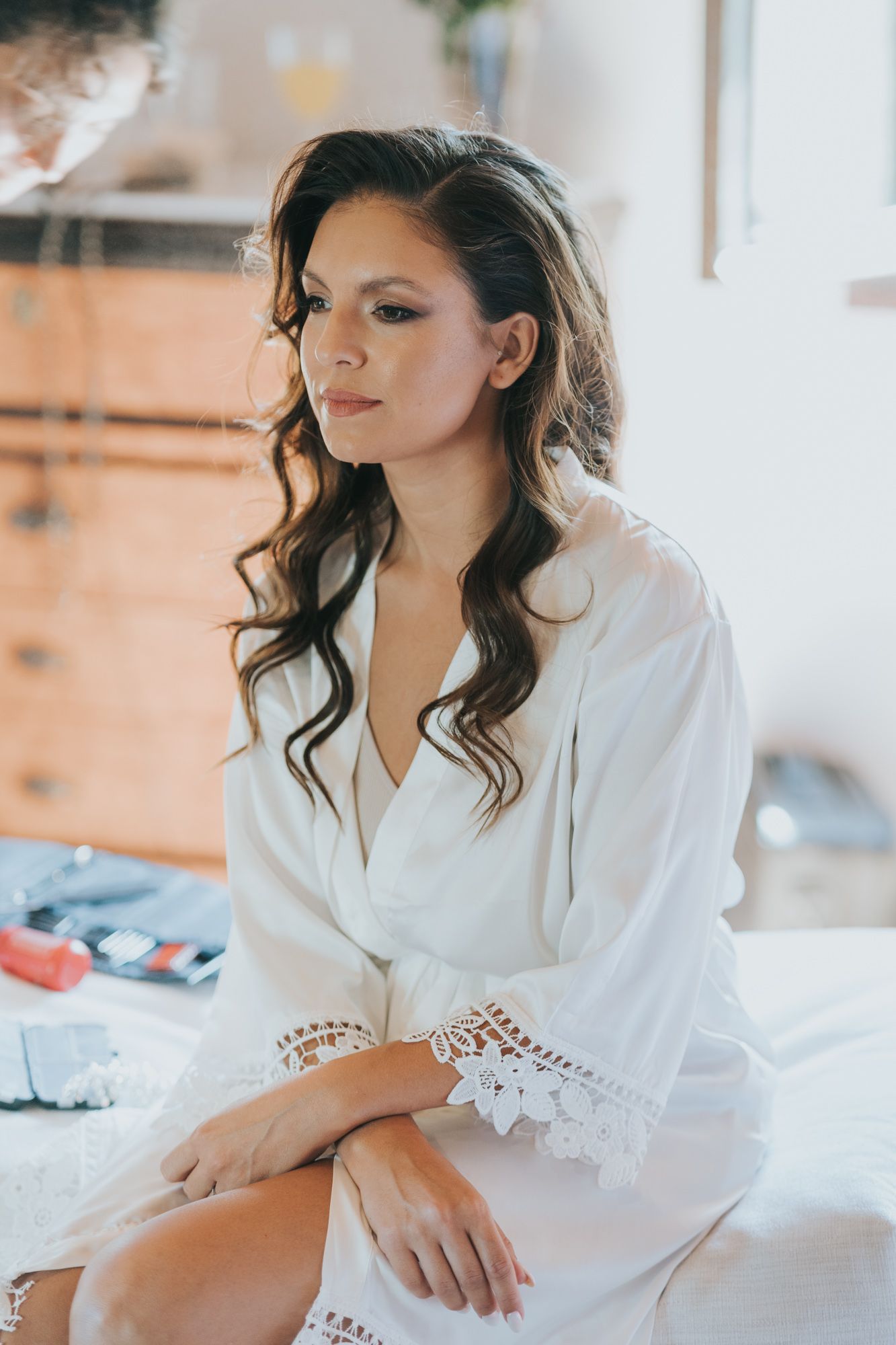 Woman in white lace robe sits on bed with long wavy dark hair and soft natural makeup in morning light.
