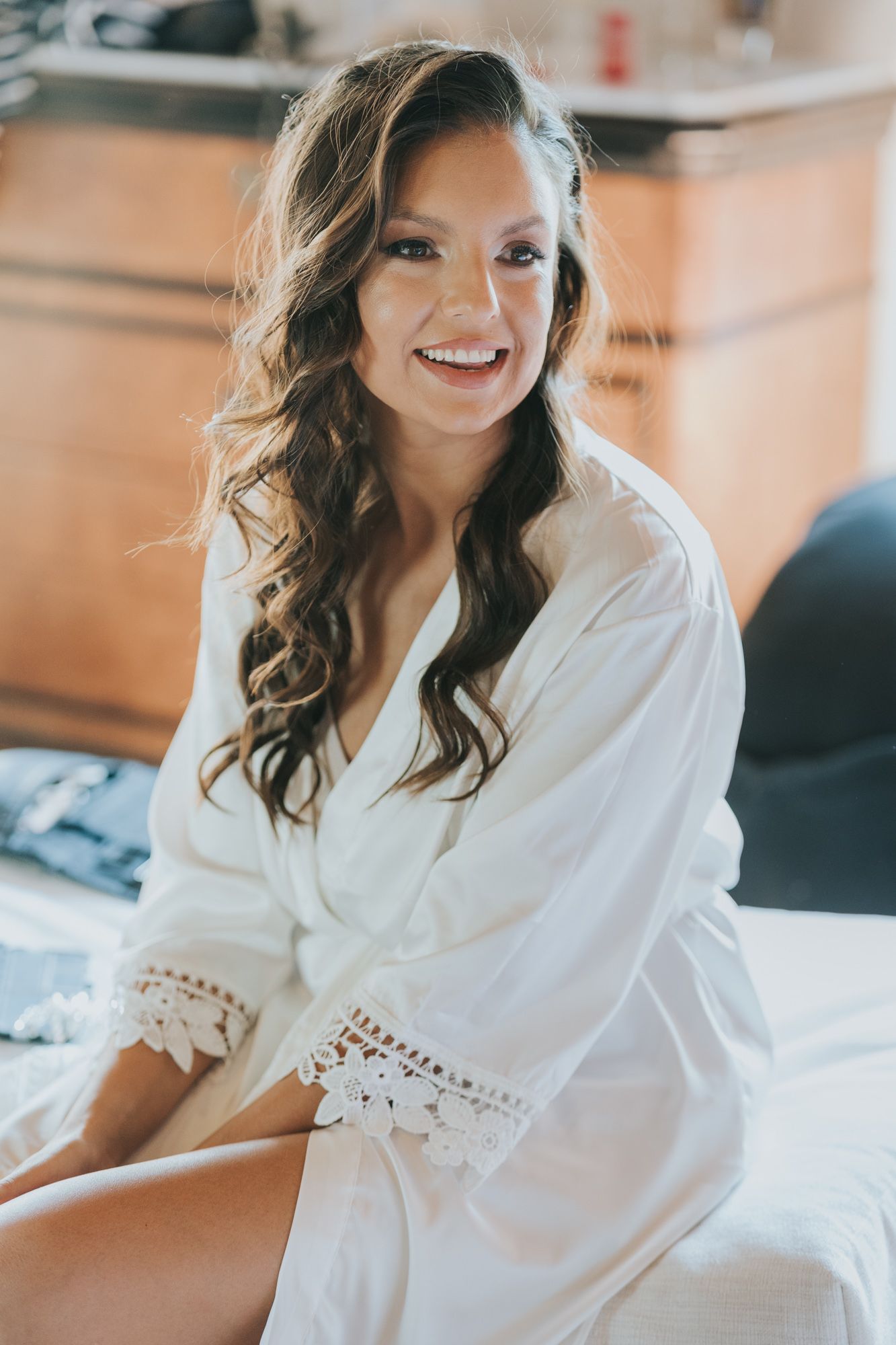 A radiant figure wearing a white lace-trimmed silk robe sits on a bed with cascading dark curls and natural lighting.
