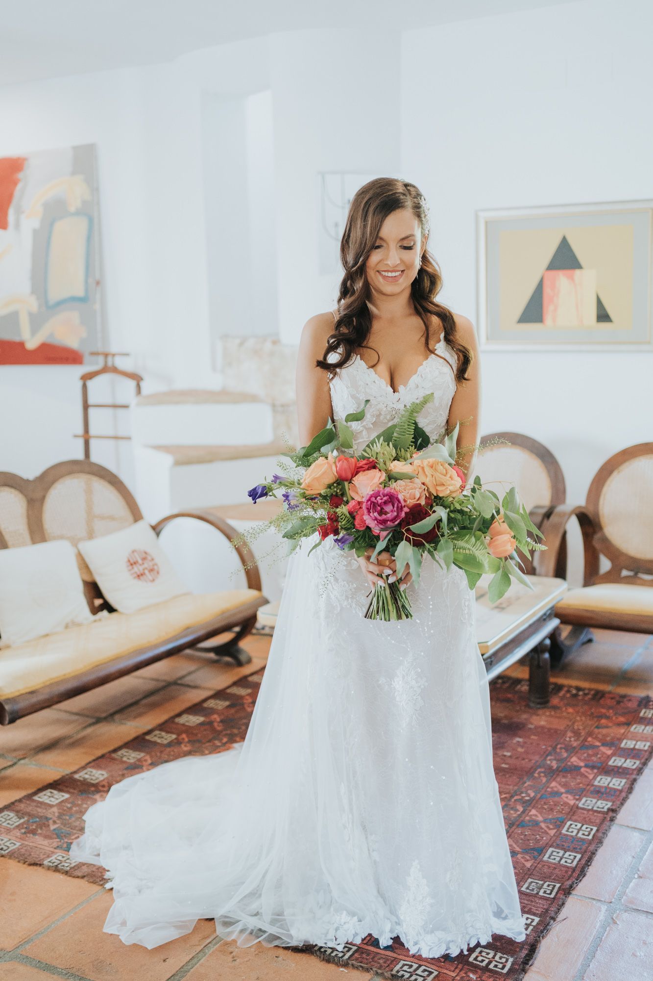 Elegant bride in lace gown holding colorful bouquet in bright modern interior.
