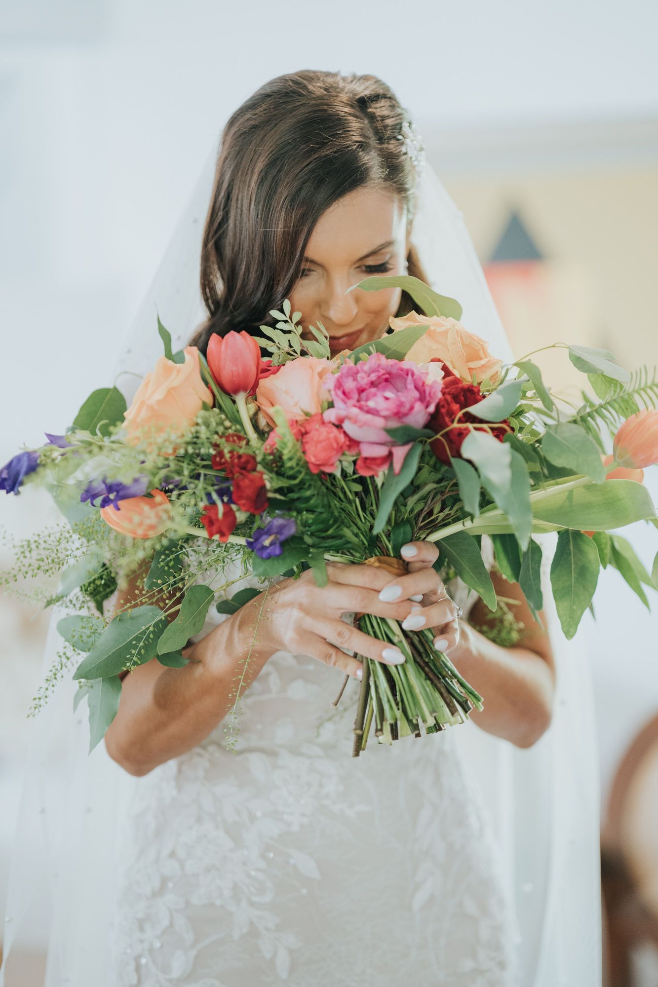 Bride in white lace dress smiles while holding vibrant bouquet of pink roses, coral flowers and greenery.