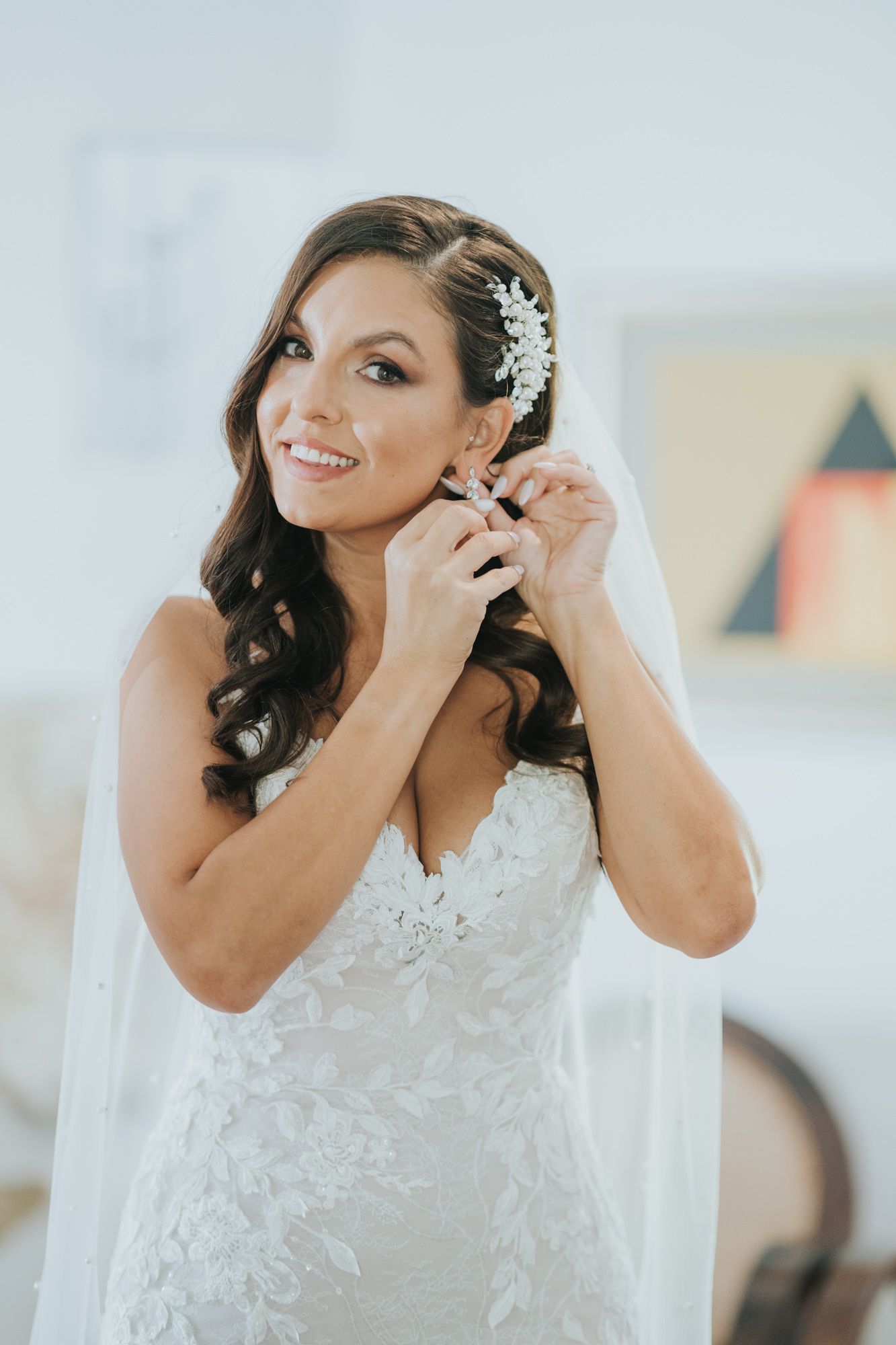 Bride in white lace wedding dress puts on earrings while wearing floral hair accessories and veil.