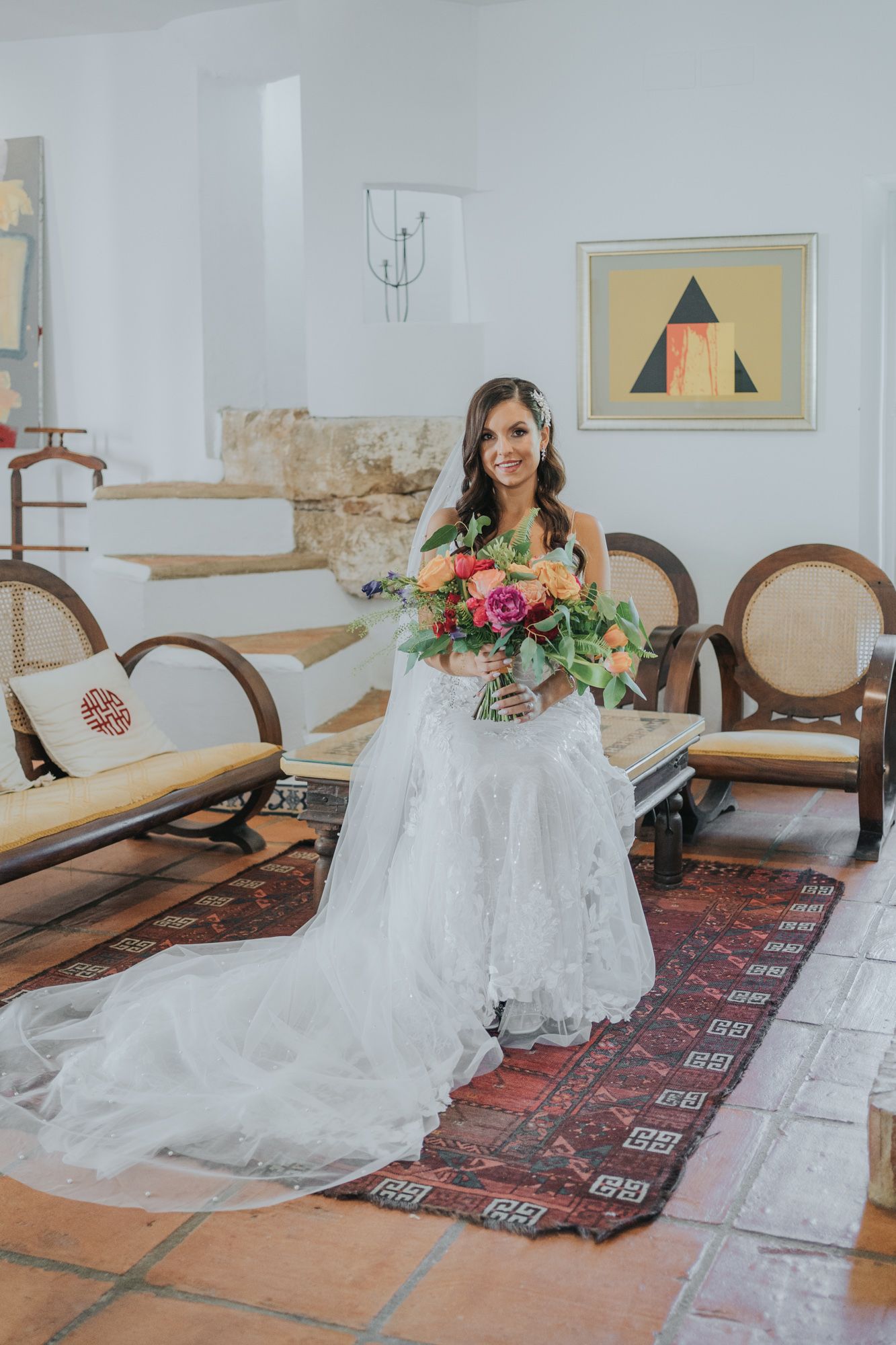 A bride poses in a white lace wedding dress with long train on a patterned rug in an elegant indoor setting.