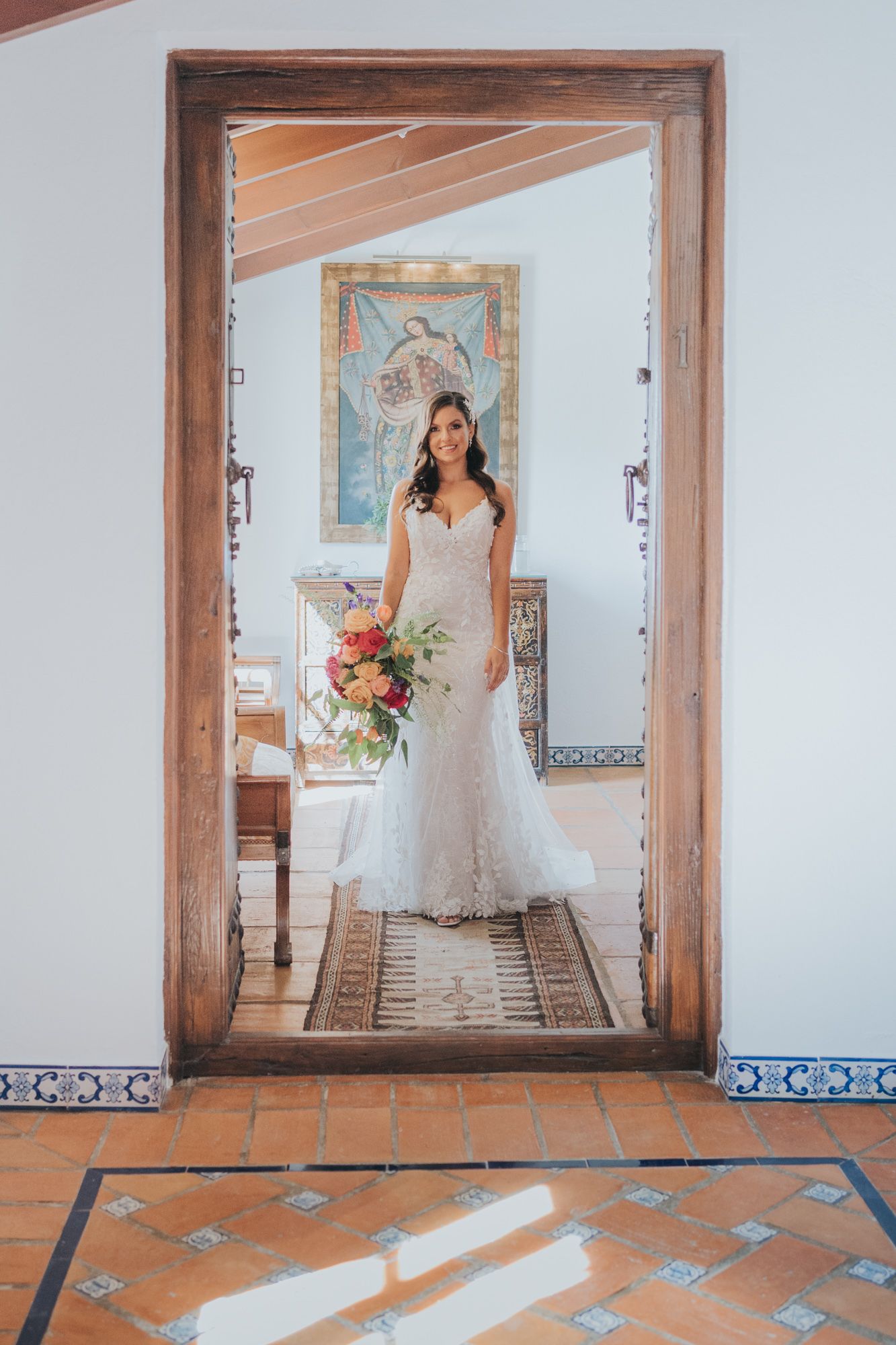 A bride in a fitted lace gown stands framed by a wooden doorway with decorative tiles and white walls.