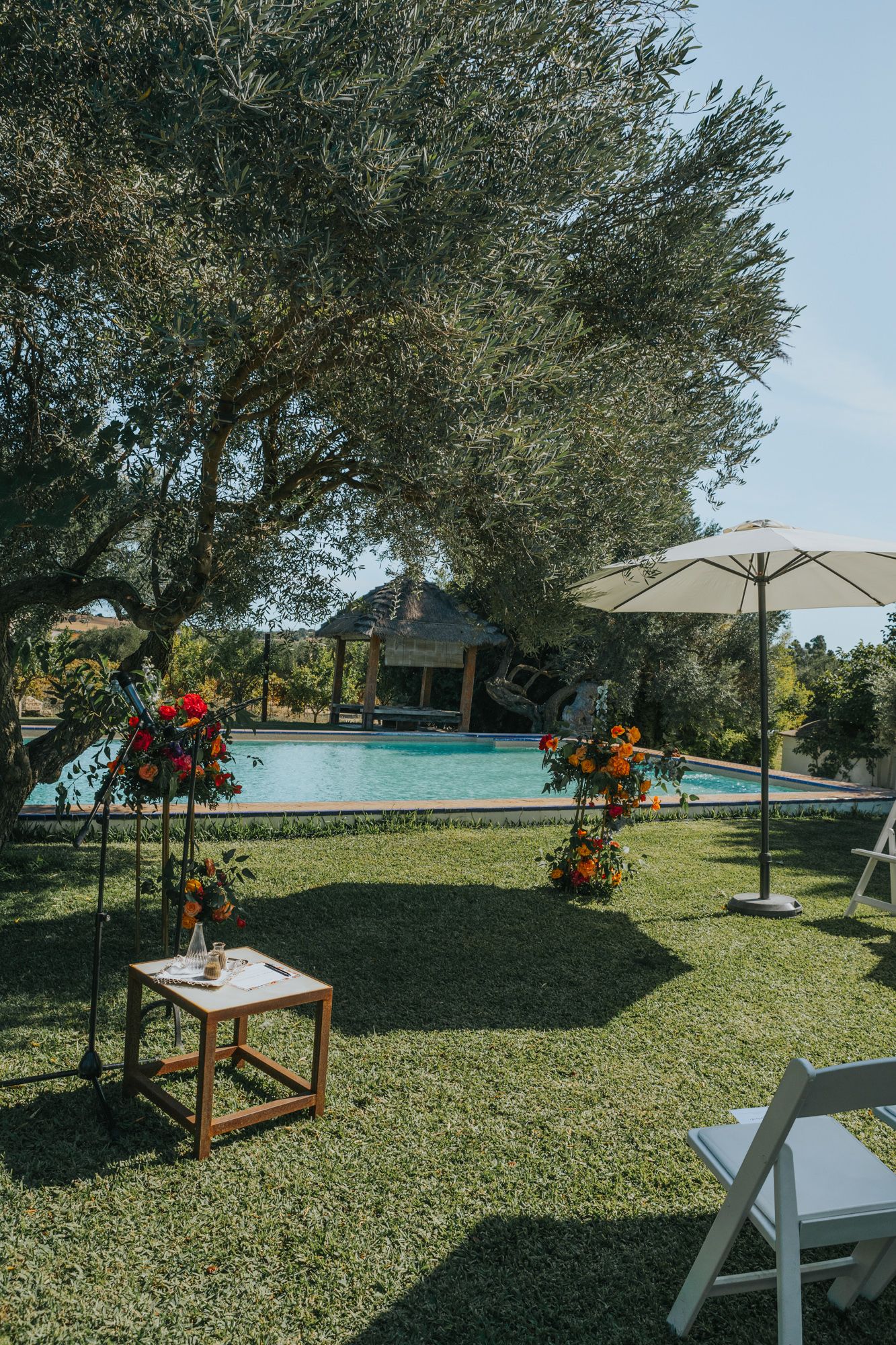 Outdoor wedding venue with swimming pool, floral arrangements, and shaded seating under olive trees.