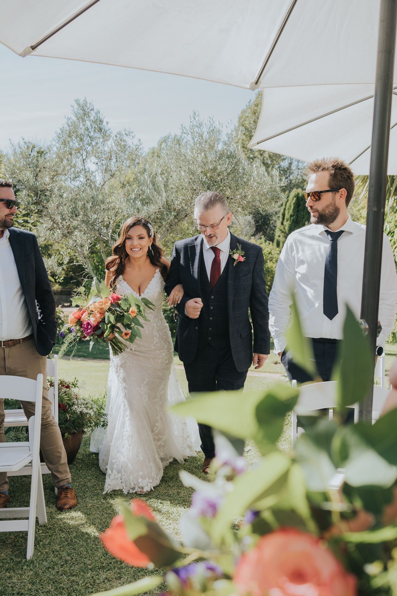 Wedding party walks under a white tent canopy during an outdoor ceremony amid olive trees and floral decorations.
