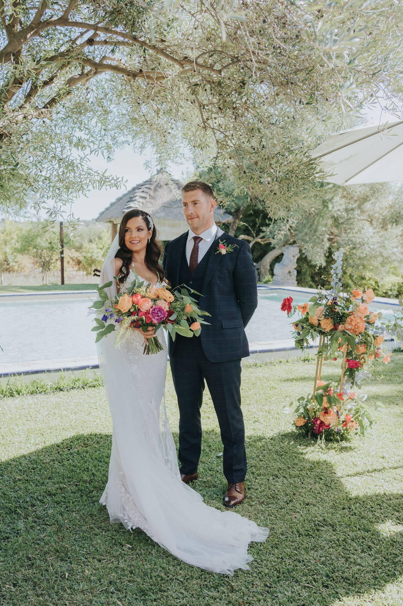 A wedding couple poses beneath an olive tree with floral decorations and greenery in an outdoor garden setting.