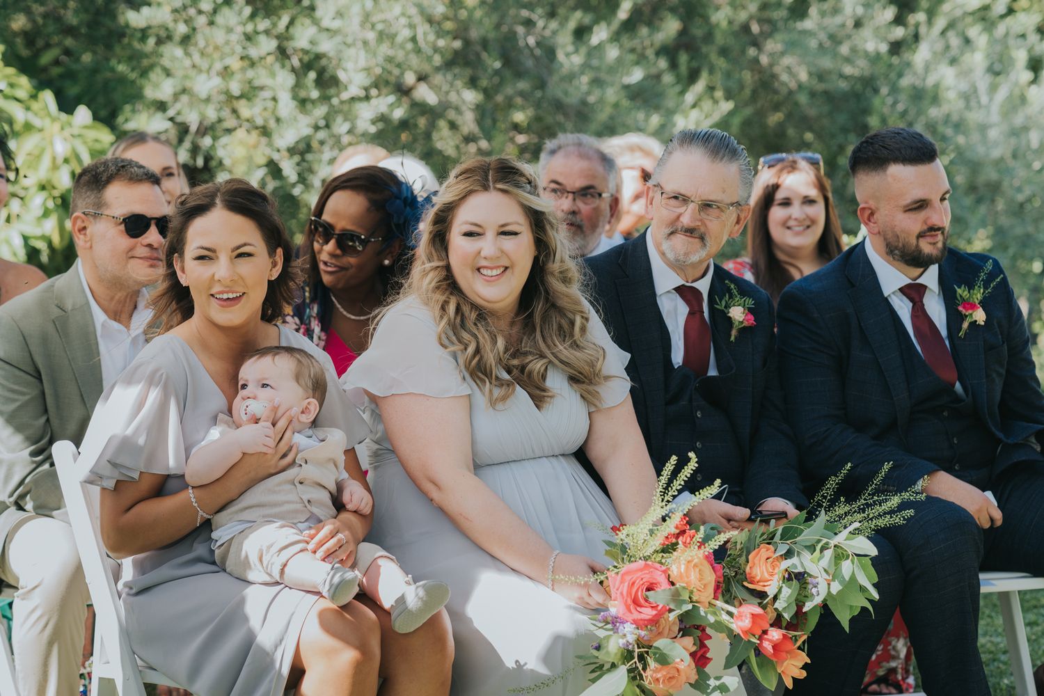 Wedding guests seated outdoors on white chairs with colorful floral arrangements.