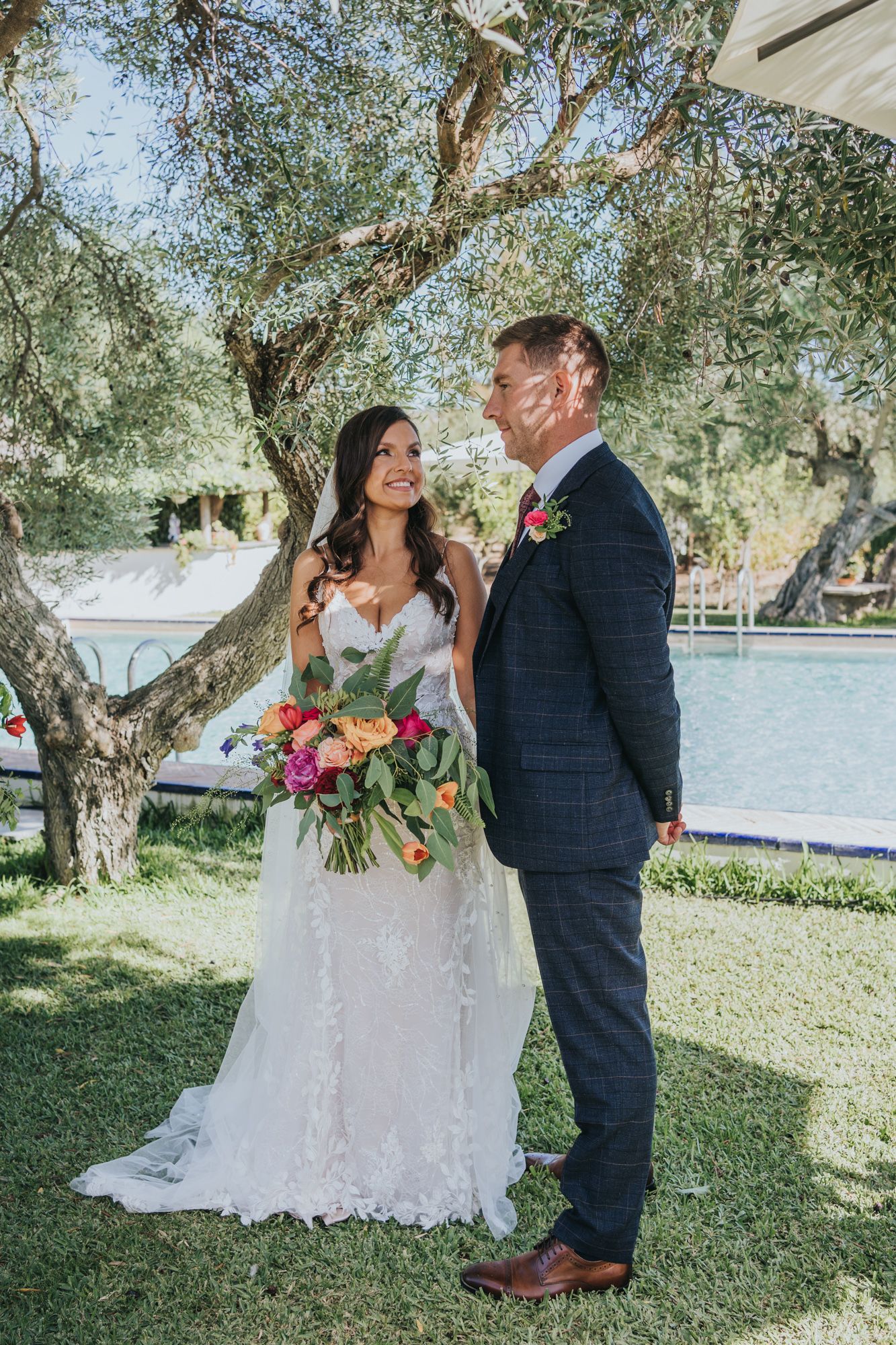 A couple in wedding attire stands together under olive trees on a grassy lawn by a waterfront venue.