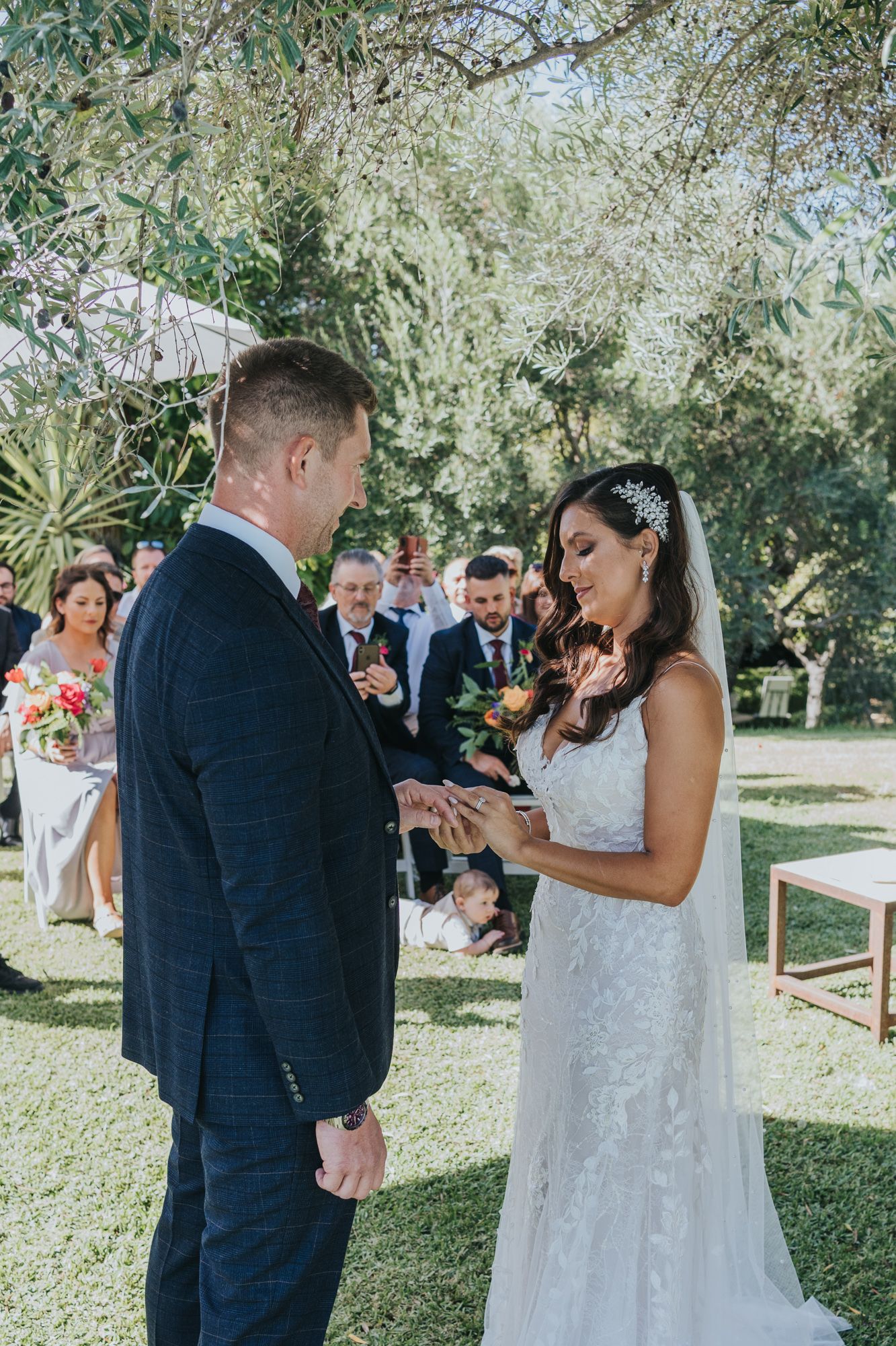 An outdoor wedding ceremony shows a couple exchanging rings under trees with guests watching.