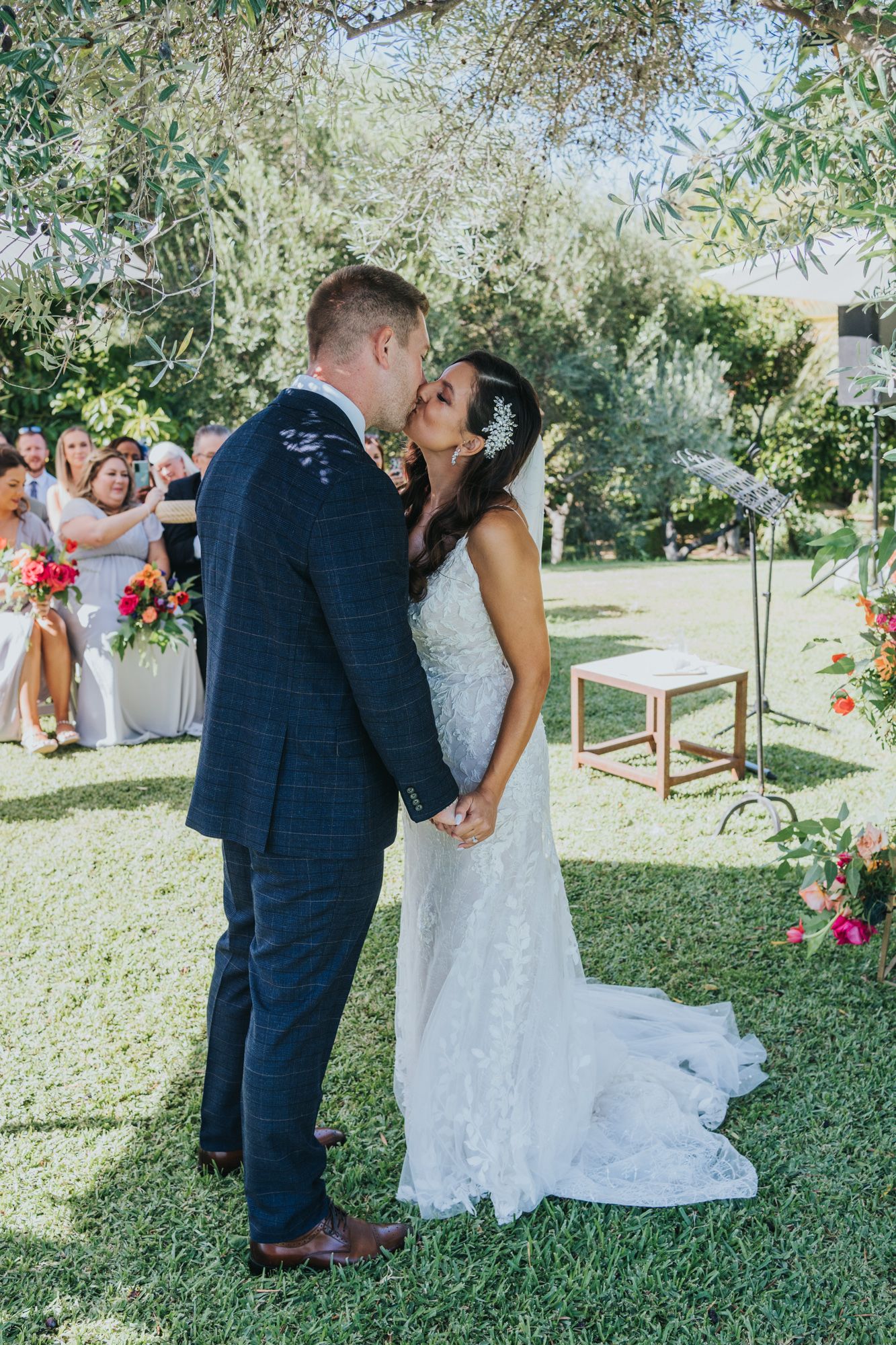 A romantic wedding ceremony kiss on a sunny day in an outdoor garden setting with floral decorations and guests watching.