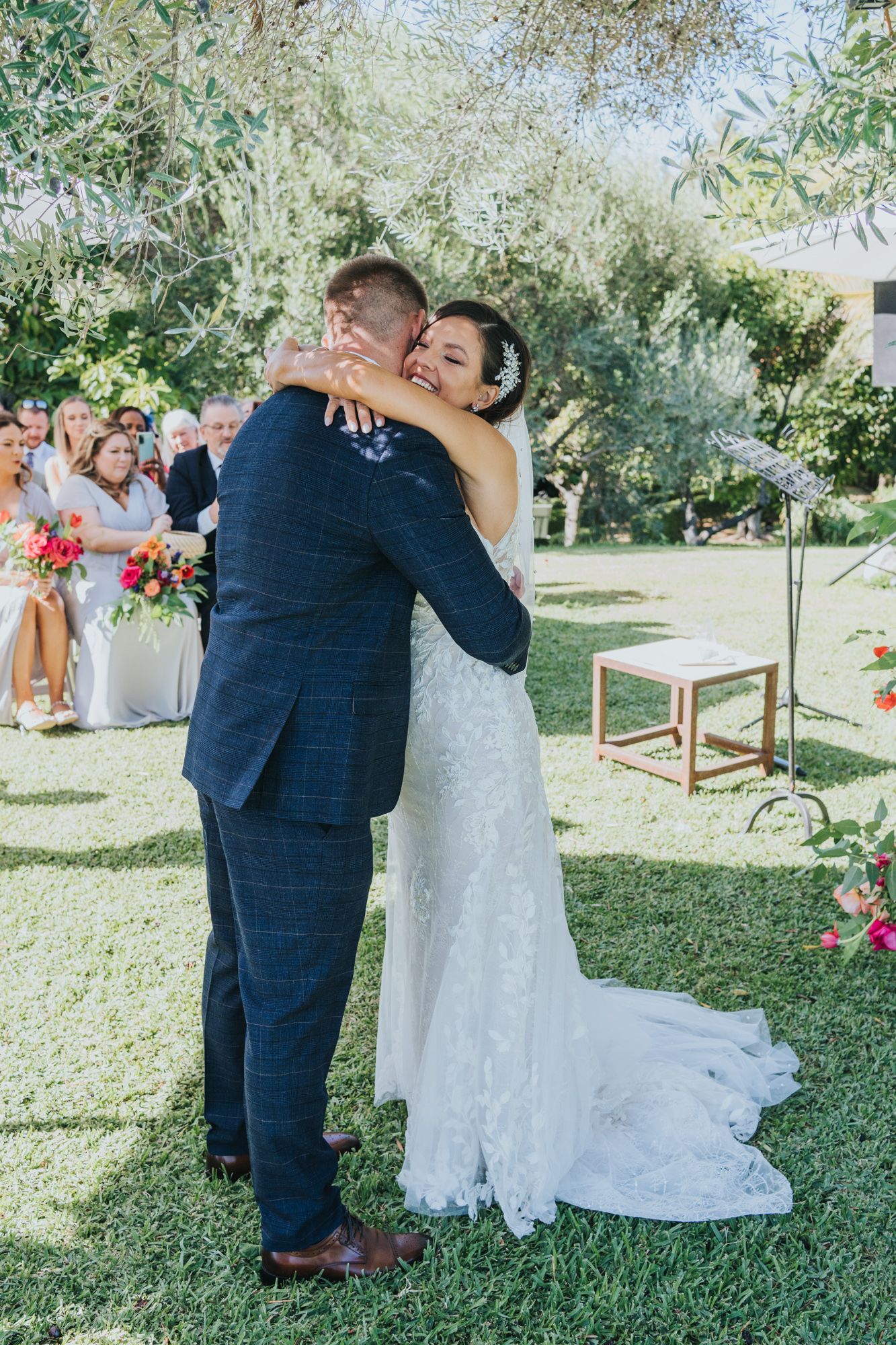 A romantic embrace between a couple in wedding attire under trees during an outdoor garden ceremony.