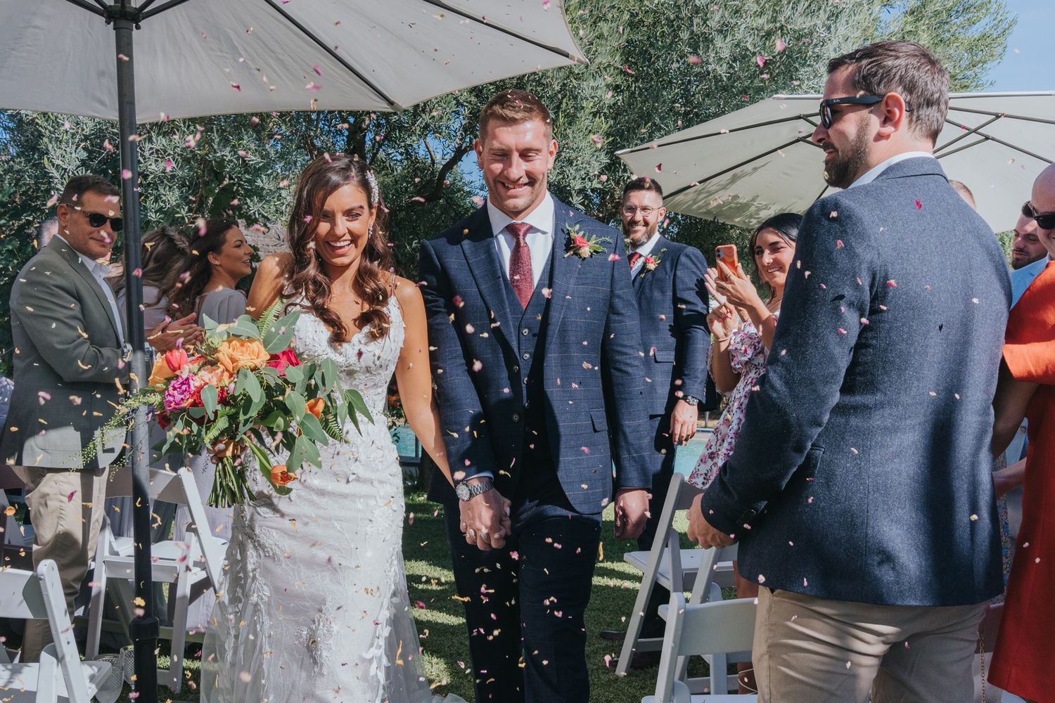 Wedding guests toss confetti over newlyweds in outdoor summer ceremony with white chairs and umbrella shade.