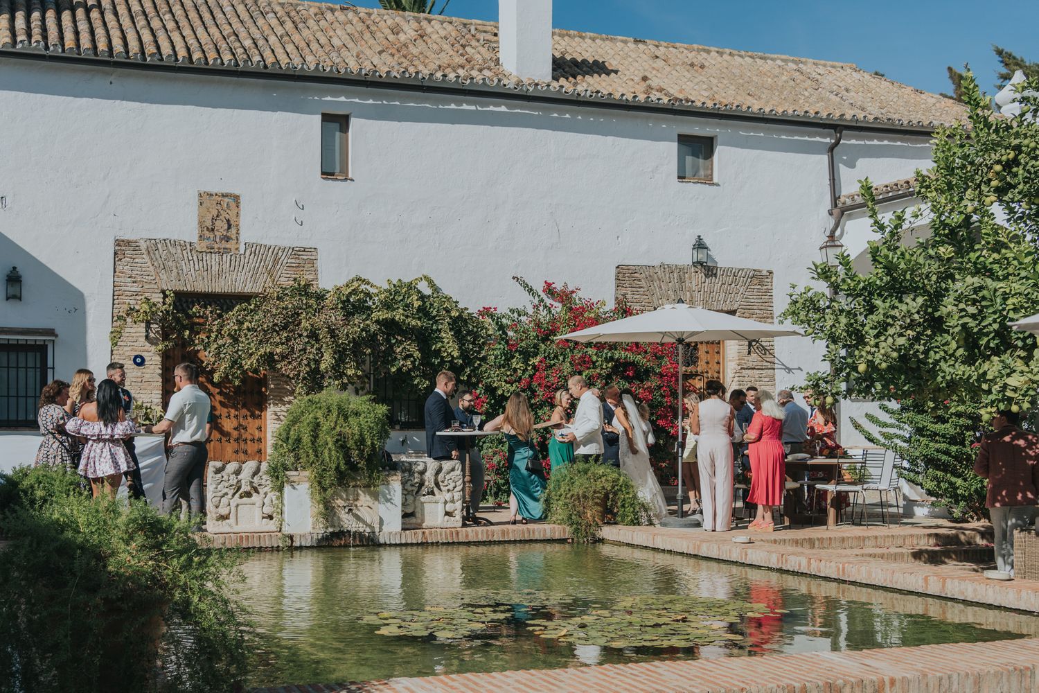 Wedding guests gathered around a courtyard pool at a Spanish-style venue during a daytime celebration.