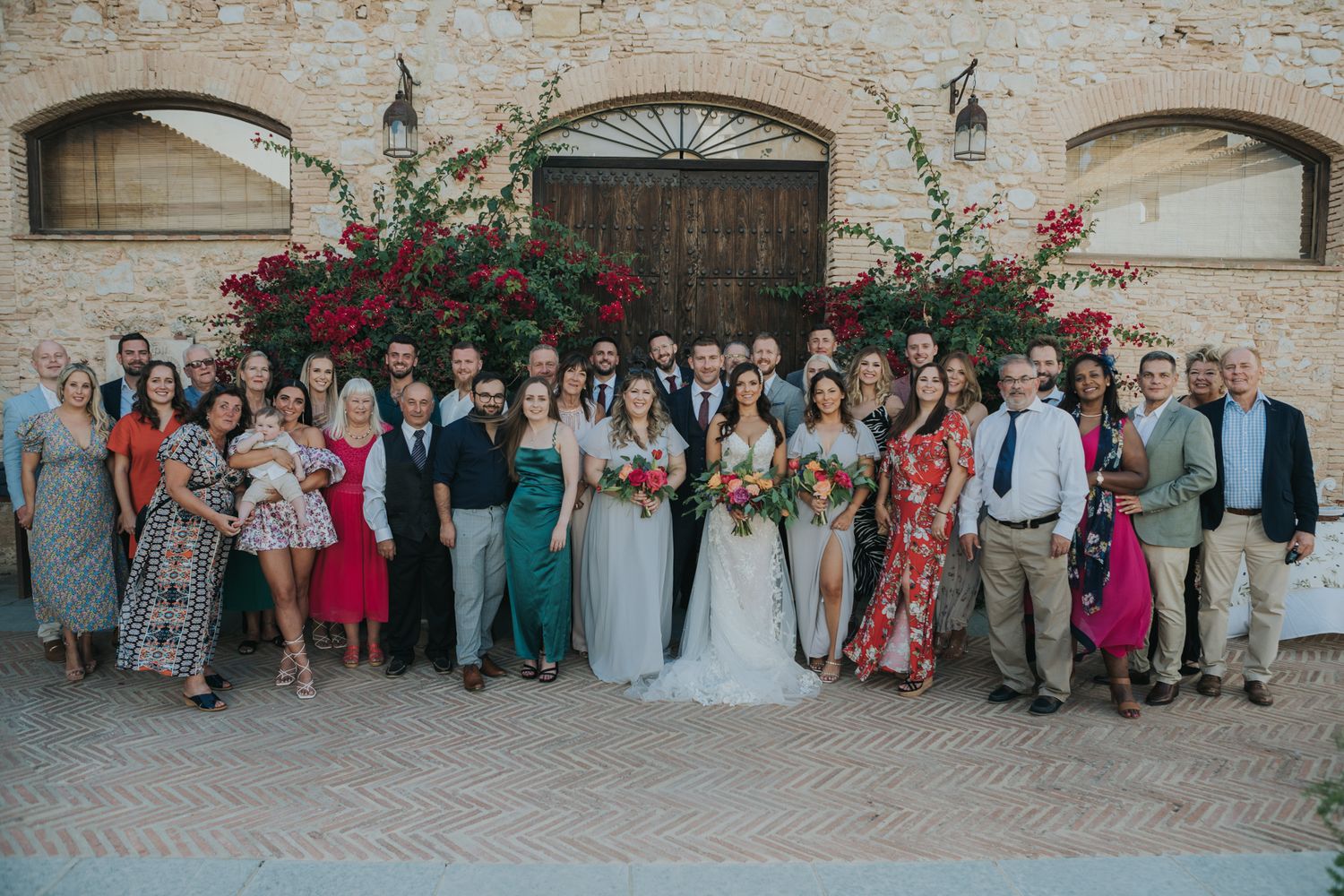 Large wedding party group photo against rustic stone wall with blooming bougainvillea.