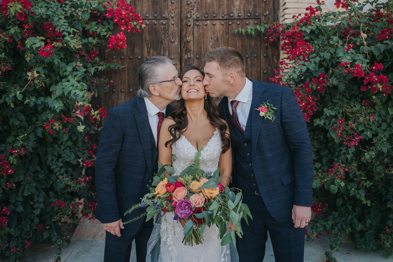 Sweet wedding moment captured in front of wooden door with blooming red flowers.