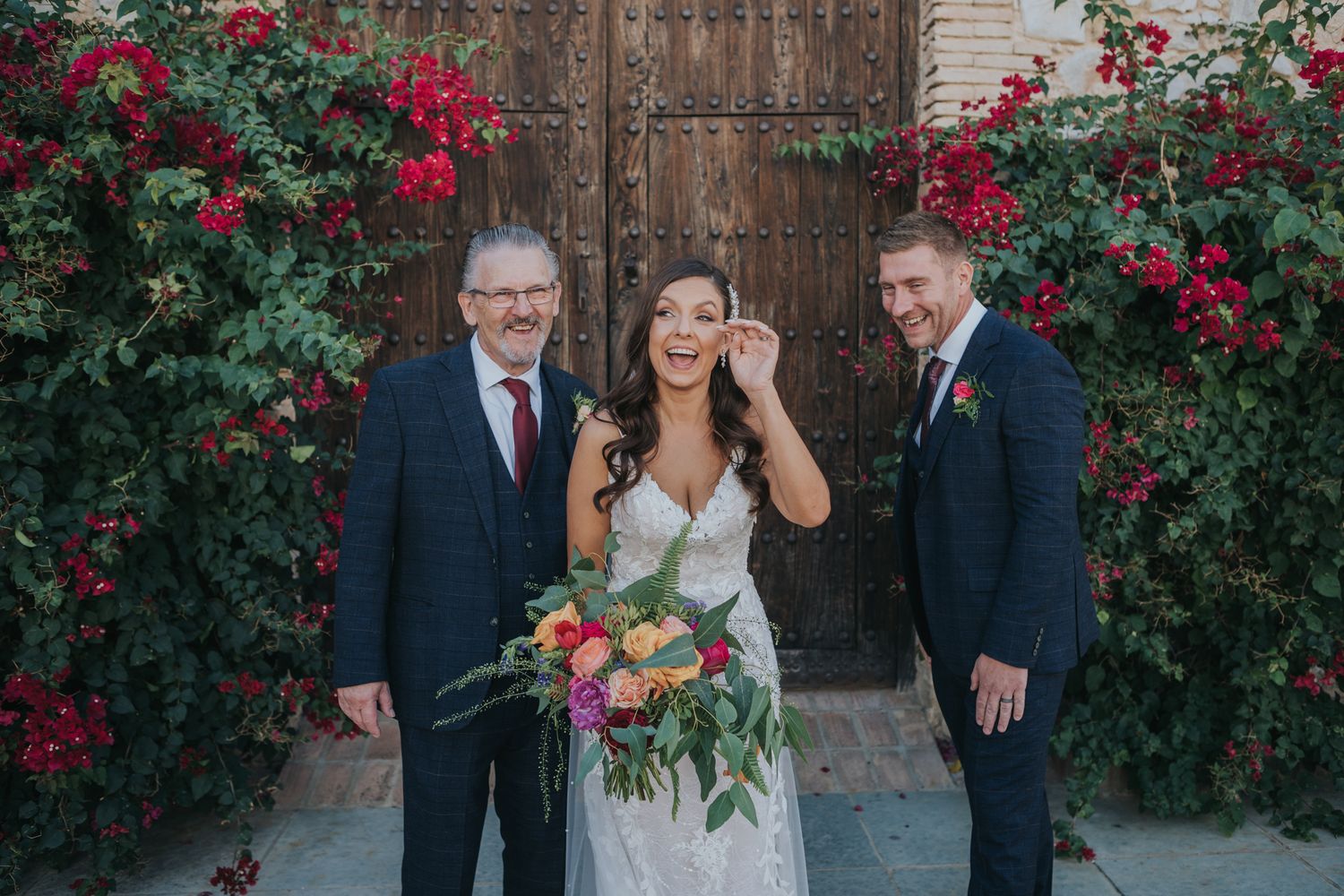 Wedding party poses together in front of a rustic wooden door surrounded by blooming red roses.