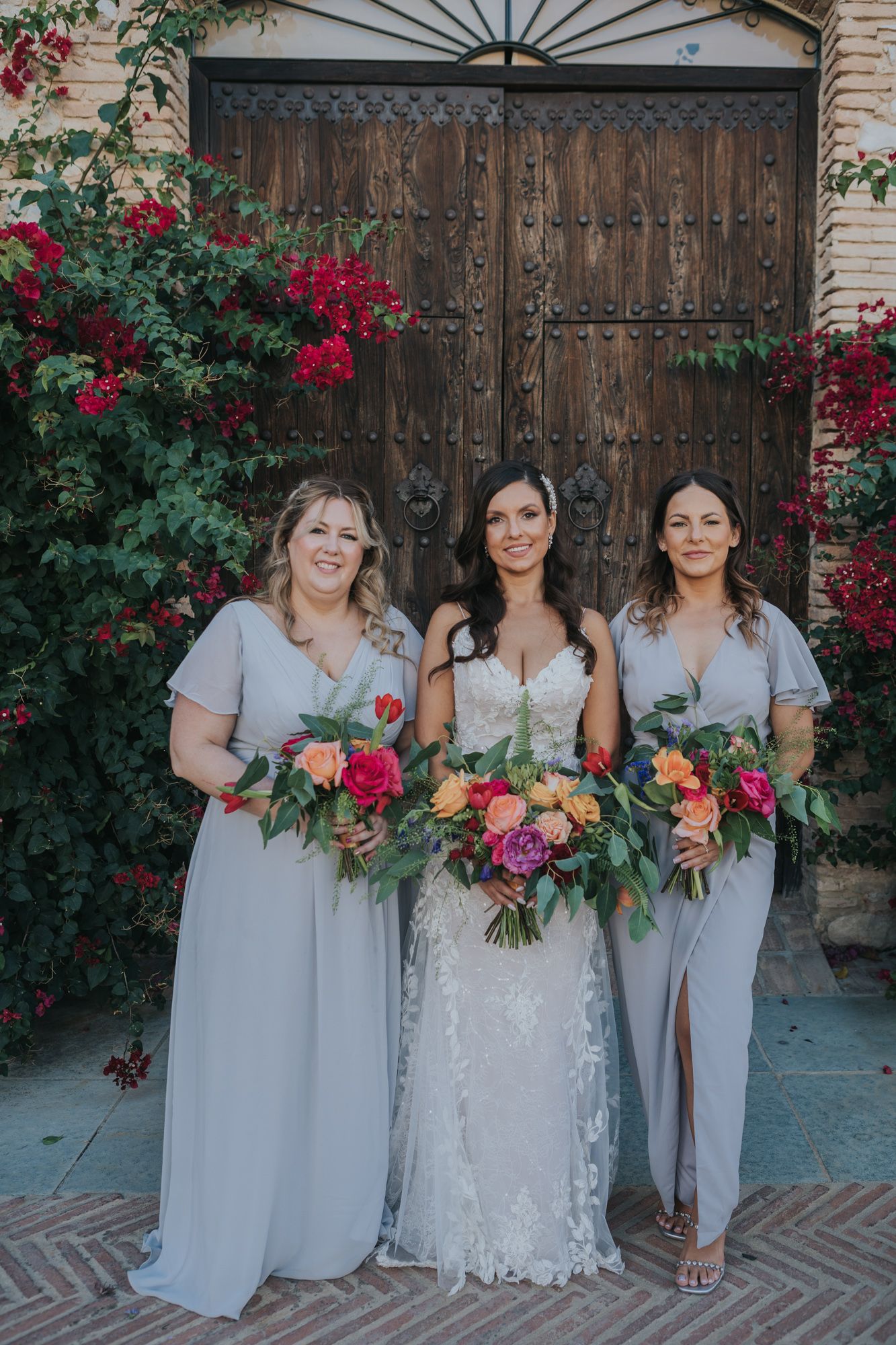 A wedding party stands in front of a rustic wooden door with vibrant red bougainvillea flowers surrounding the entrance.