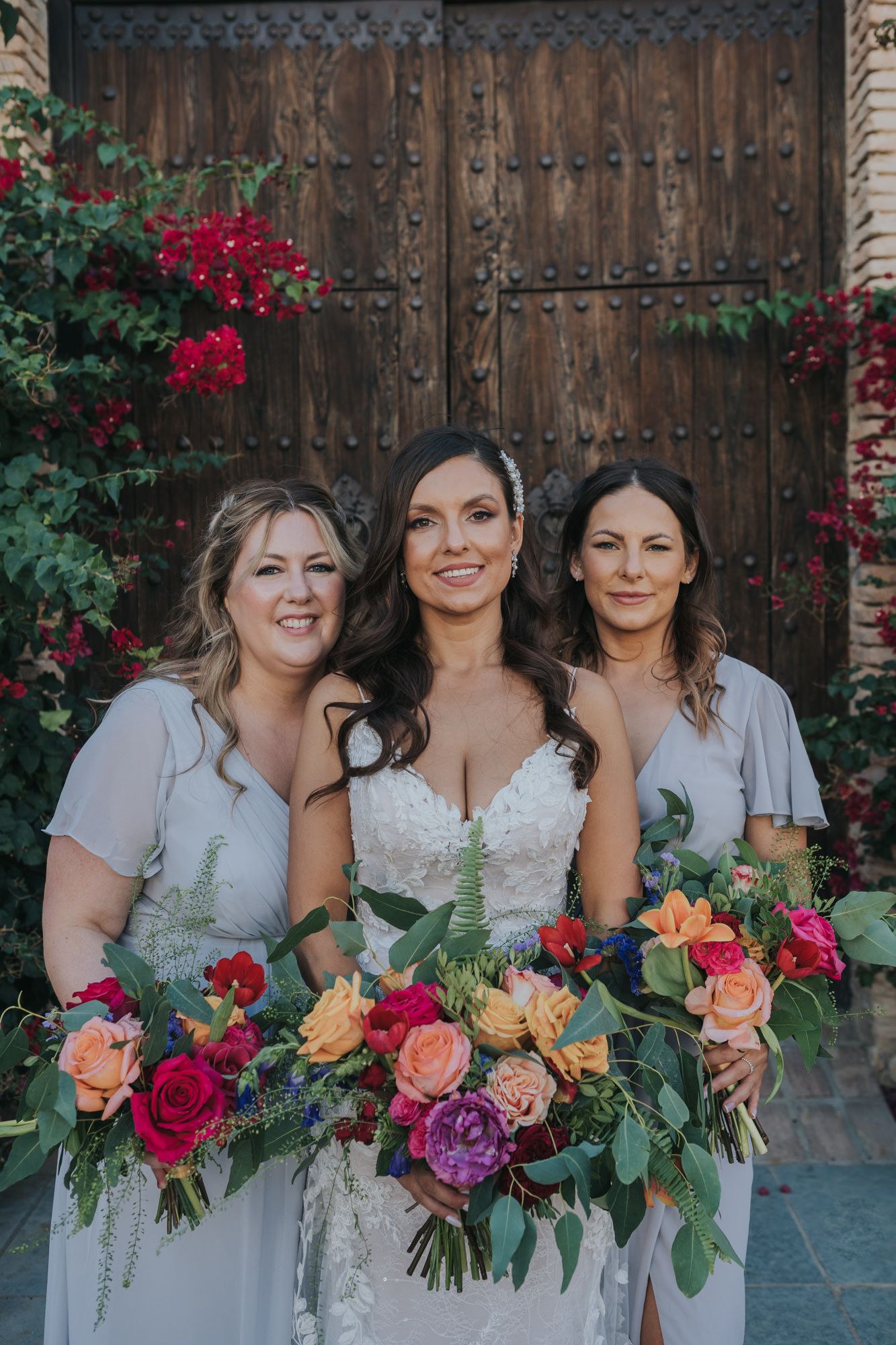 Three women stand together holding vibrant floral bouquets in front of a rustic wooden door with red bougainvillea.