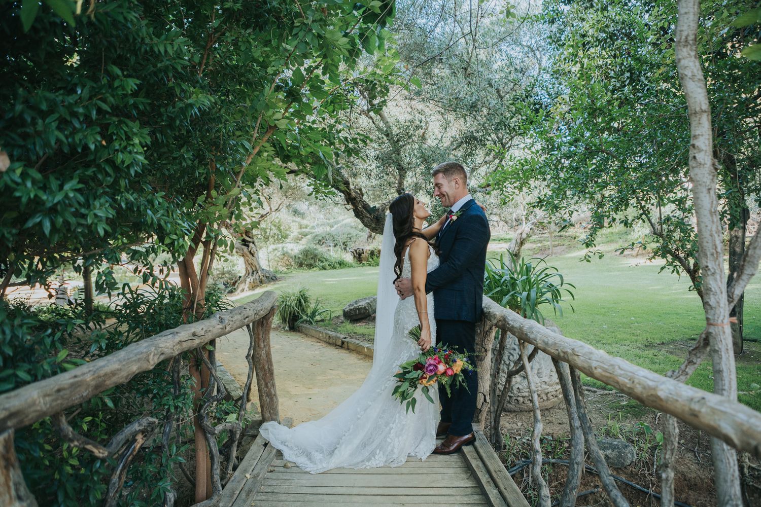 Newlyweds sharing a romantic moment on a rustic wooden bridge surrounded by lush trees.