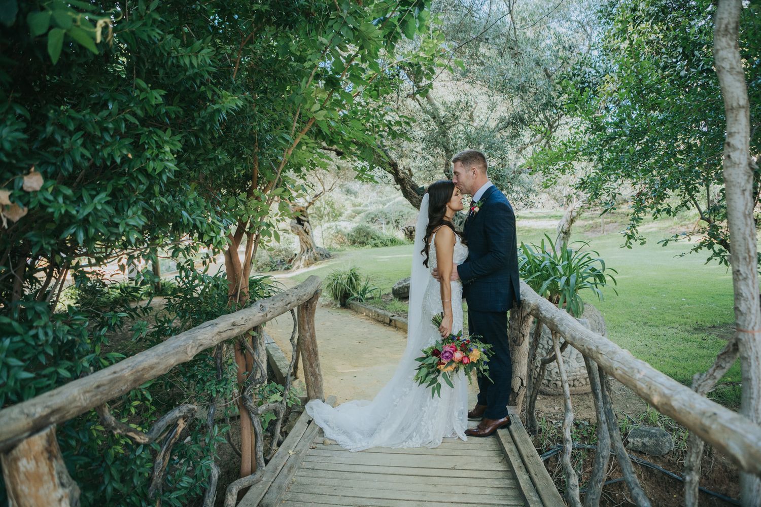 A romantic wedding photo of a couple embracing on a rustic wooden bridge surrounded by lush greenery and trees.