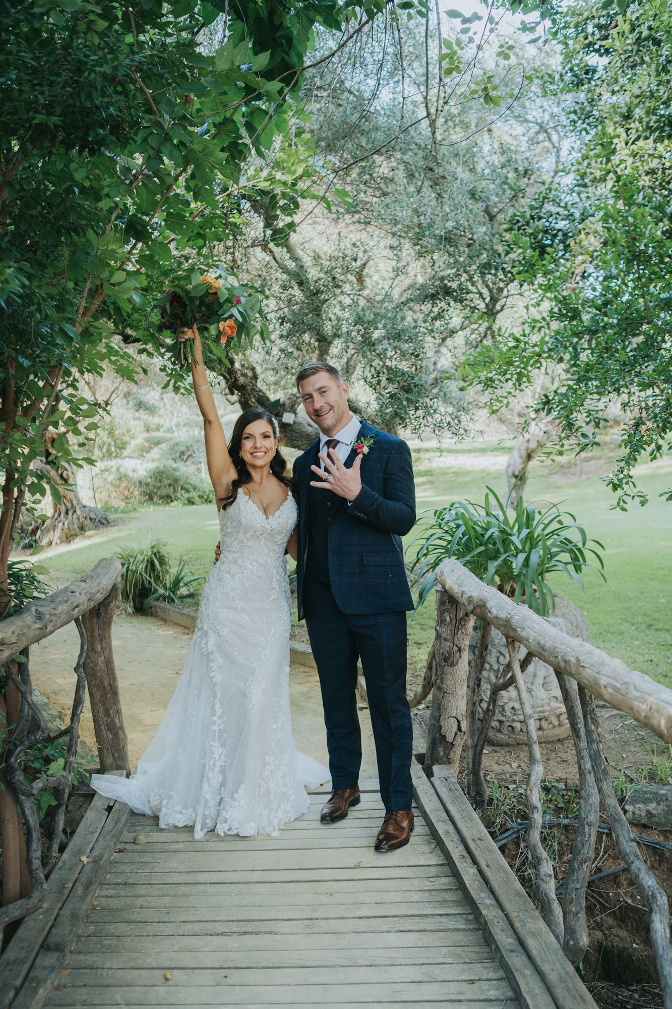 A couple celebrates on a rustic wooden bridge surrounded by lush greenery in a garden setting.