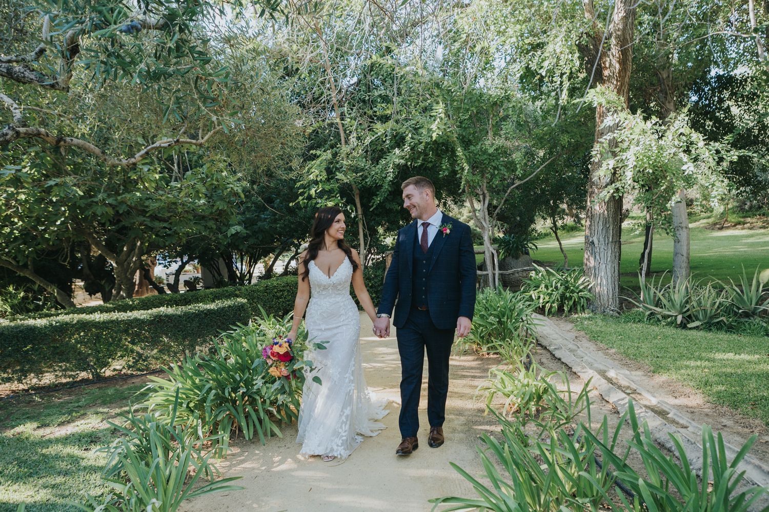 Couple walking along a garden path surrounded by greenery and flowering plants.