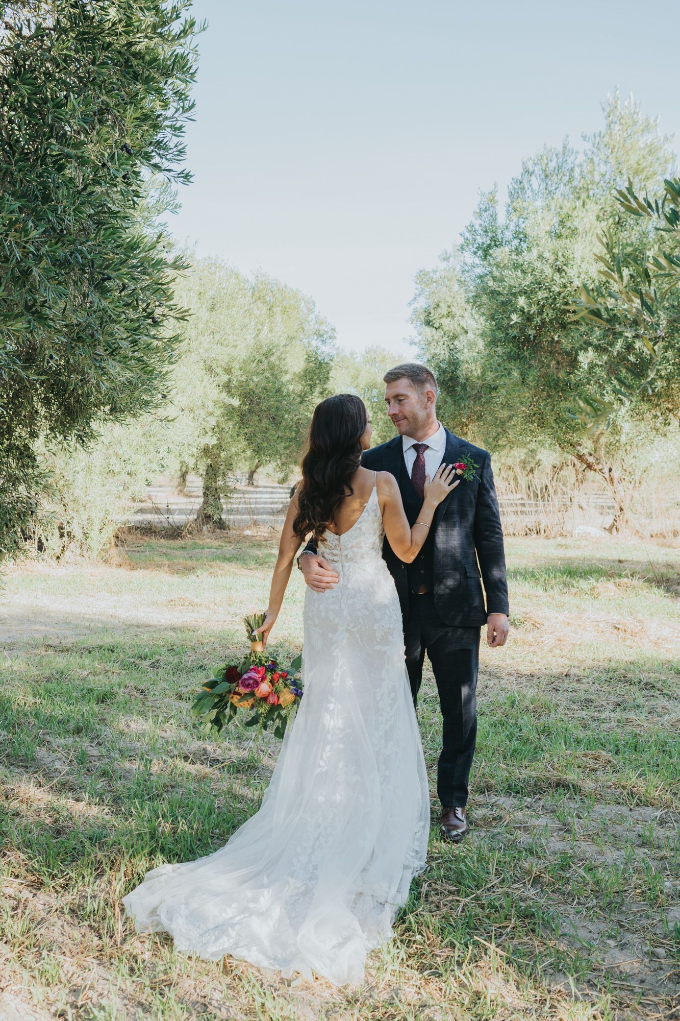 Bride and groom pose together in an olive grove with flowing white dress and colorful bouquet.