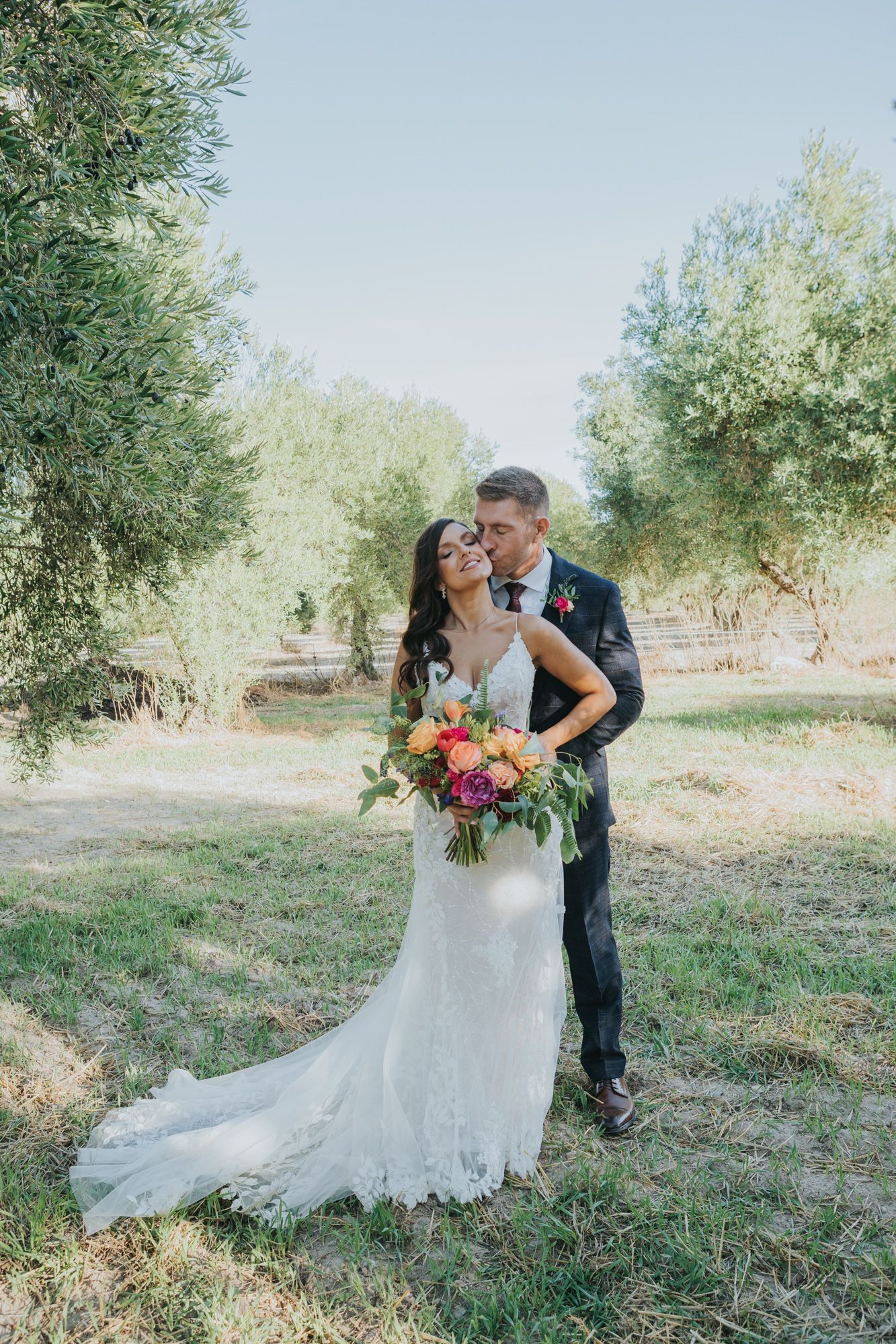 Newlyweds share a romantic moment among olive trees, with the bride holding a vibrant bouquet in her lace gown.