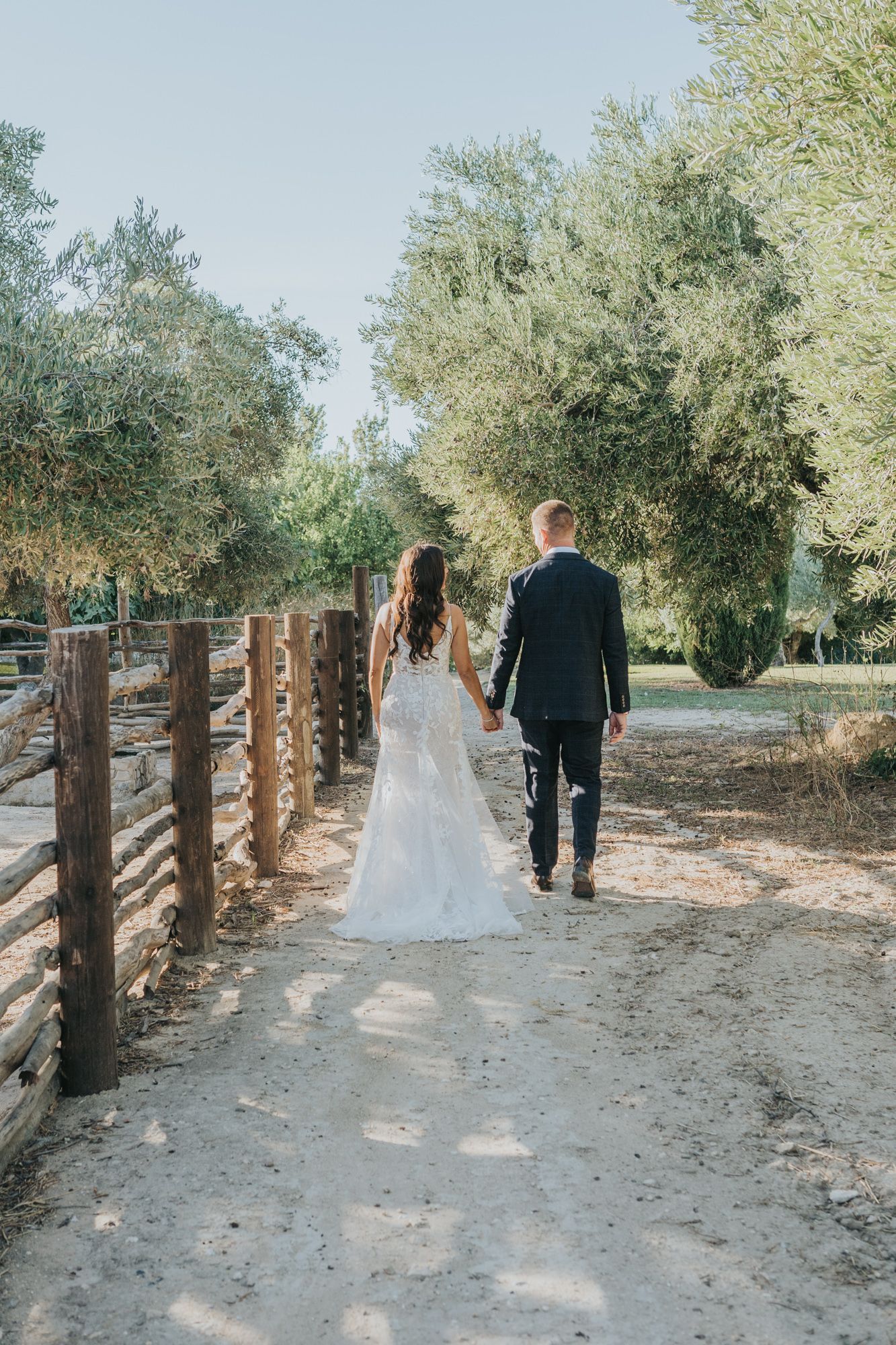 Newlyweds walking down a rustic path lined with wooden fencing and olive trees.