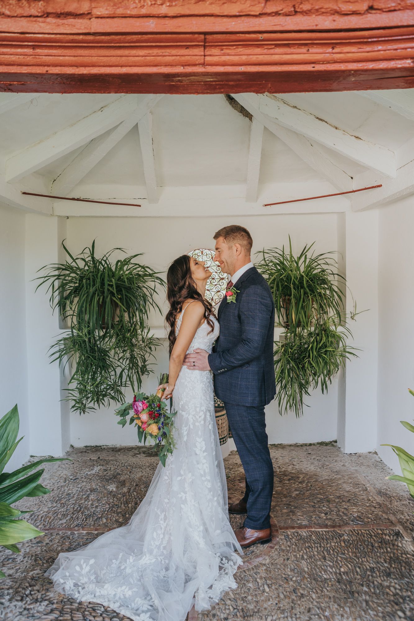 A beautiful wedding moment captured in a white gazebo with hanging plants as newlyweds embrace in elegant attire.