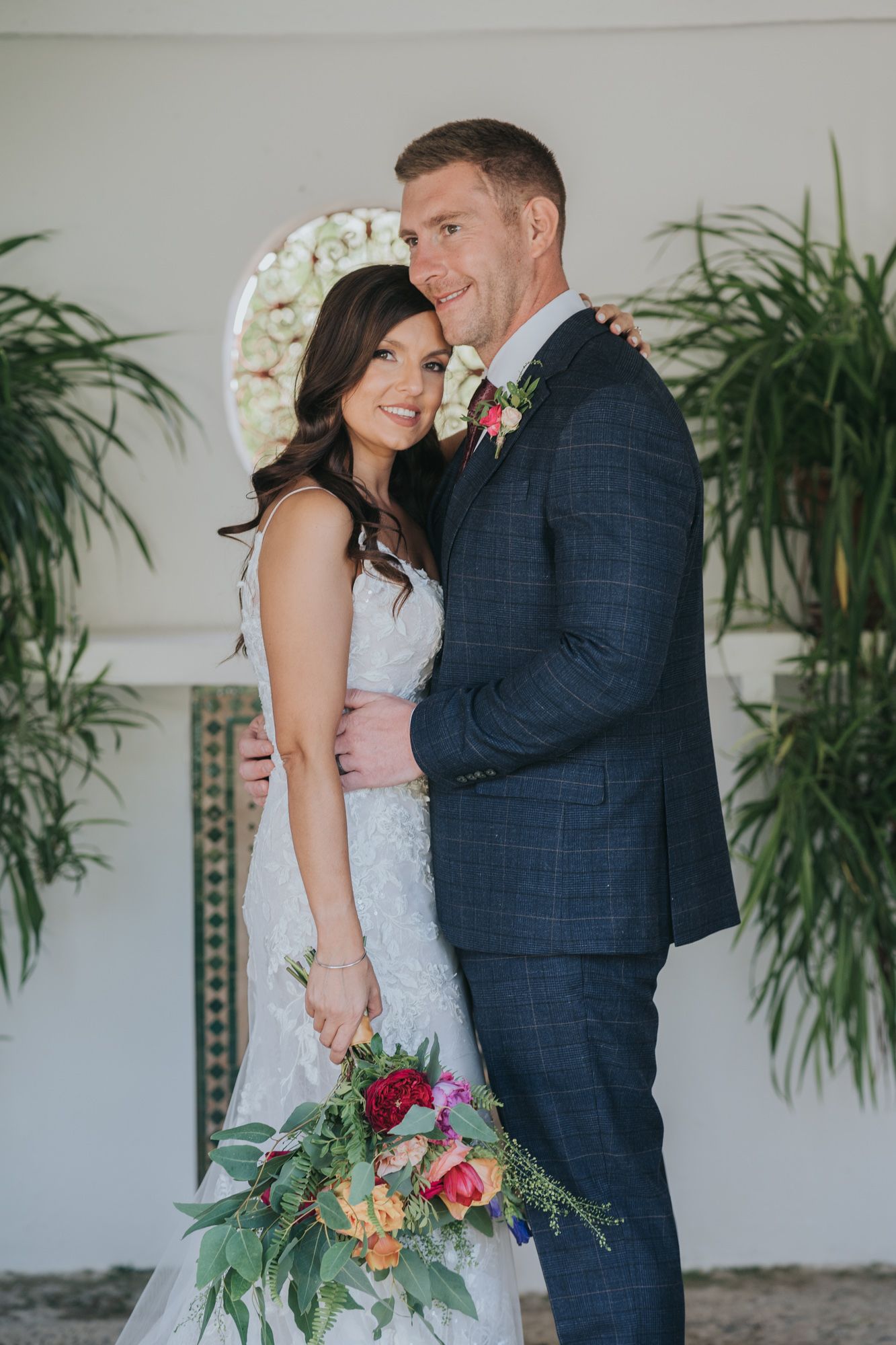 Elegant wedding couple posing together indoors with tropical plants and a decorative arch backdrop.