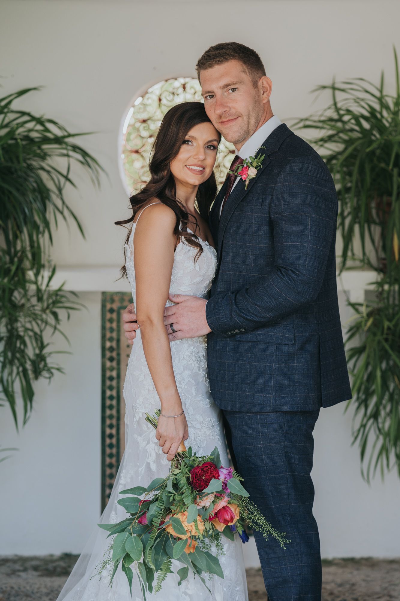 Wedding couple in formal attire pose with colorful bouquet against white wall with tropical plants.