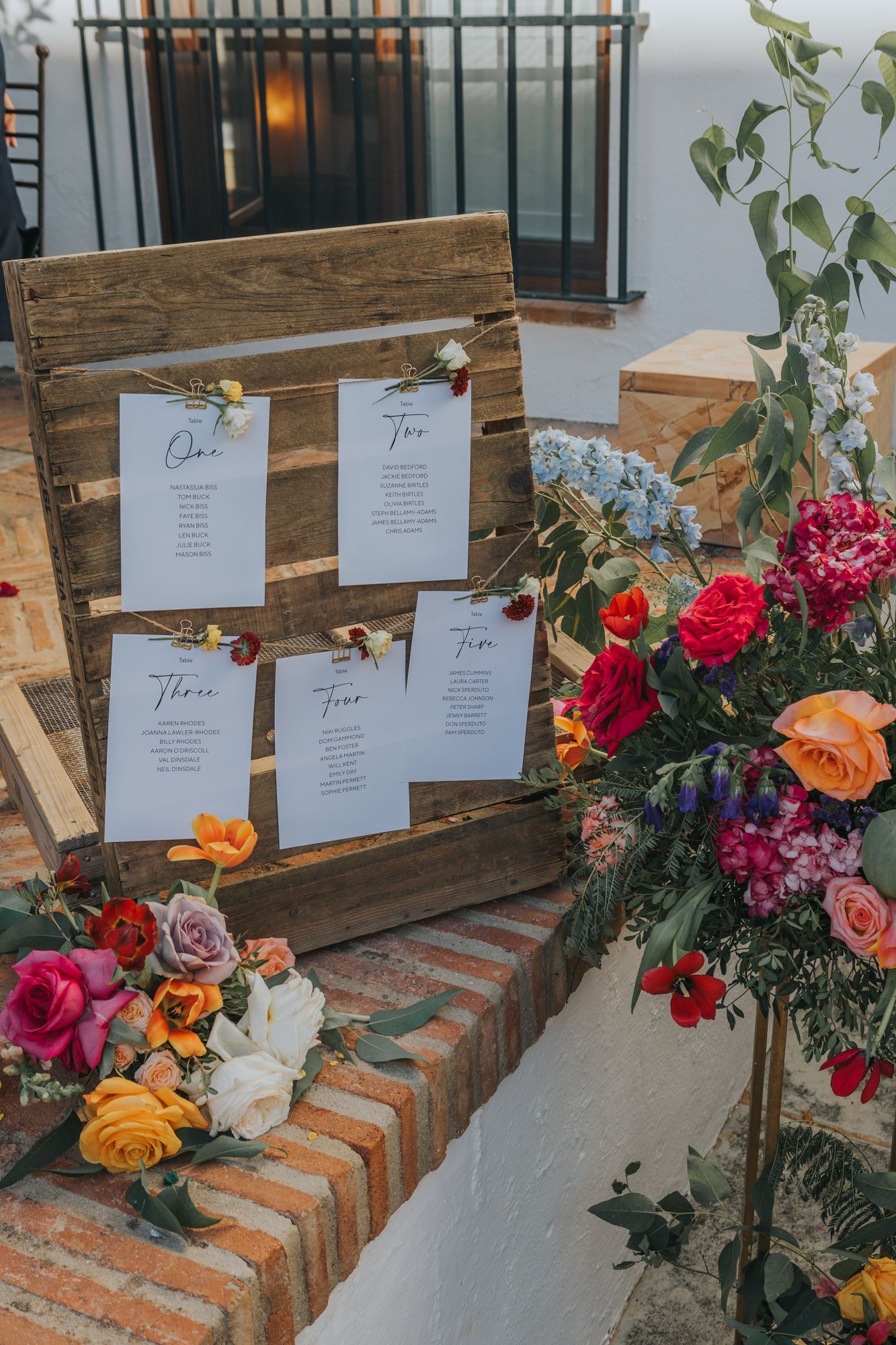 Wedding seating chart on wooden board with colorful floral arrangements featuring roses and mixed blooms.