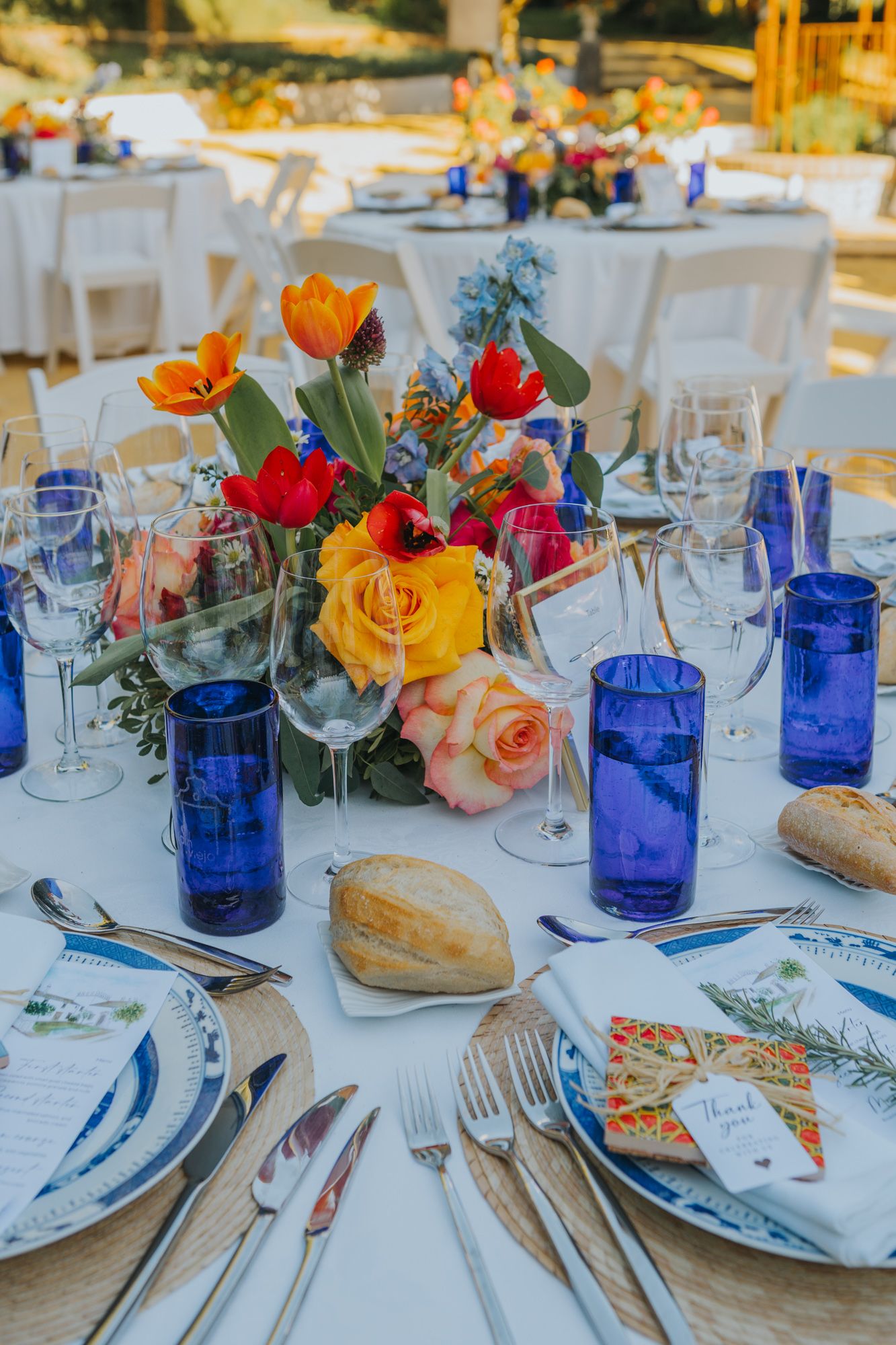 Elegant wedding table setting with colorful floral centerpiece, blue glassware, and place settings on white linens.