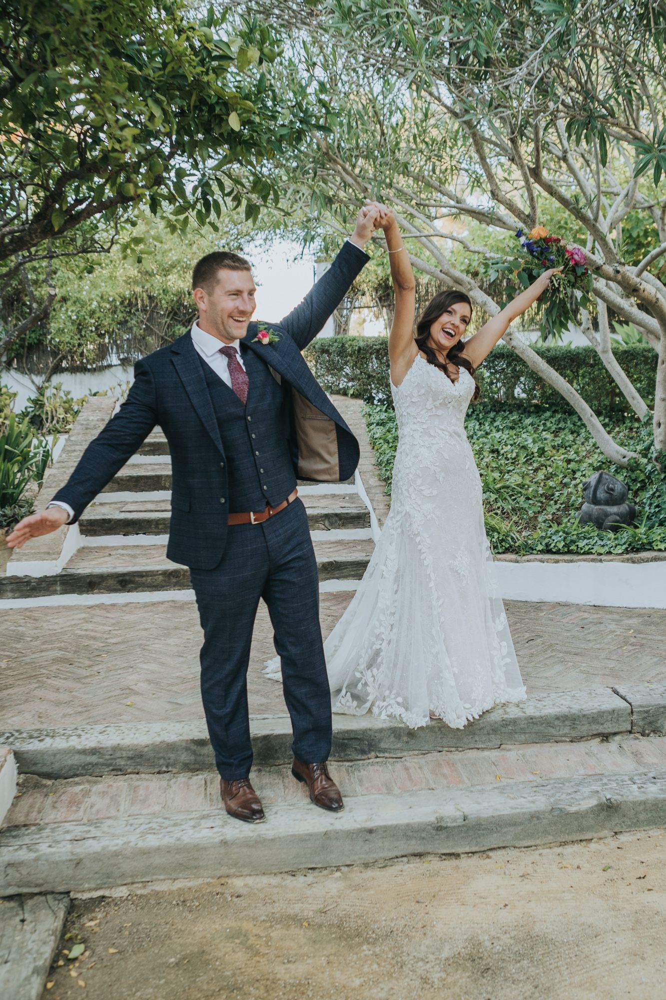 A couple celebrates joyfully with raised arms on stone steps surrounded by lush greenery on their wedding day.