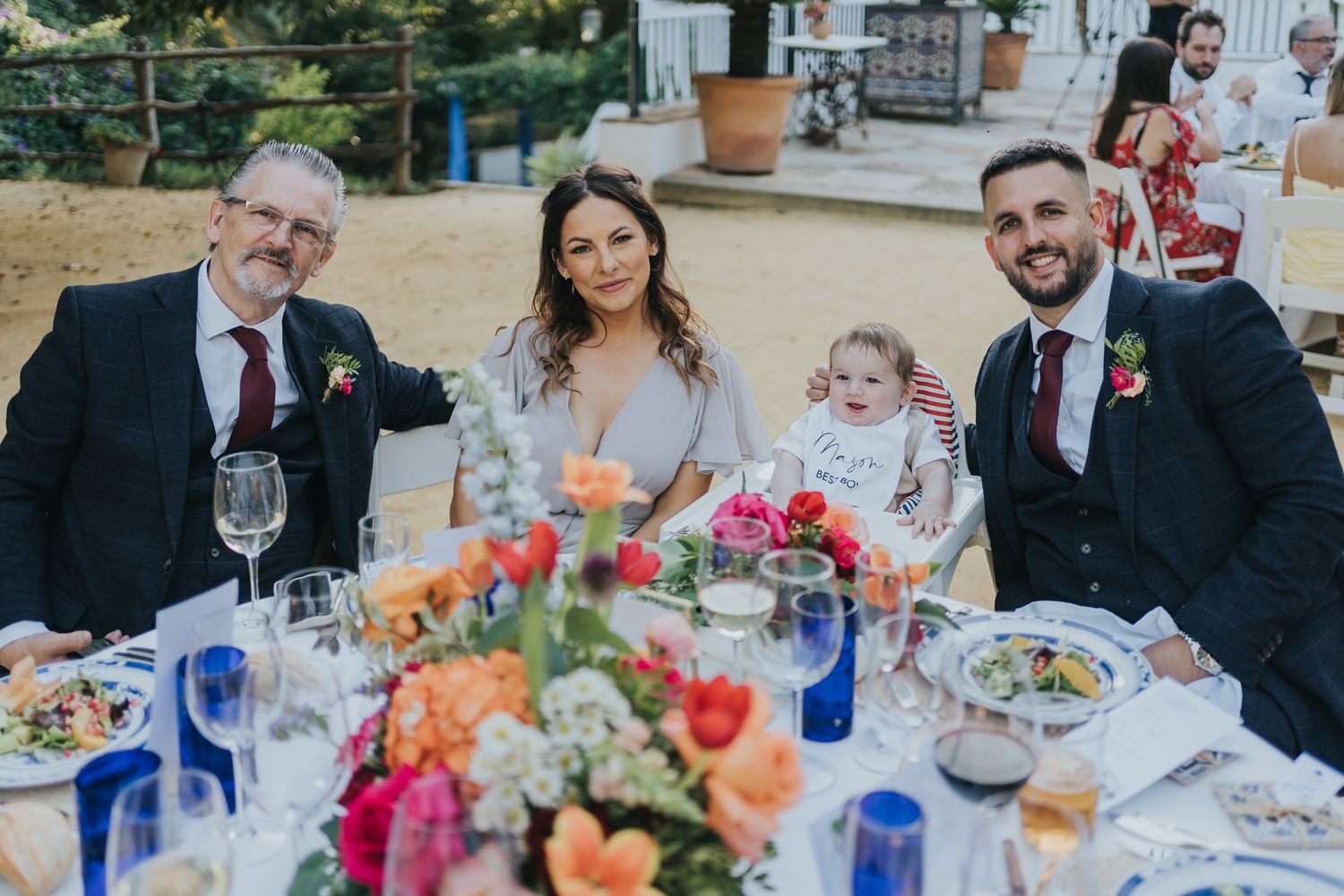 Wedding reception table with colorful floral centerpieces and family enjoying outdoor celebration.