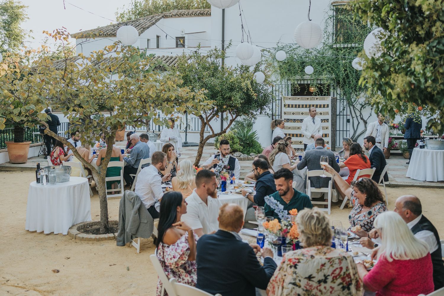 Wedding guests enjoy an outdoor reception under trees at rustic tables with white linens and floral decor.