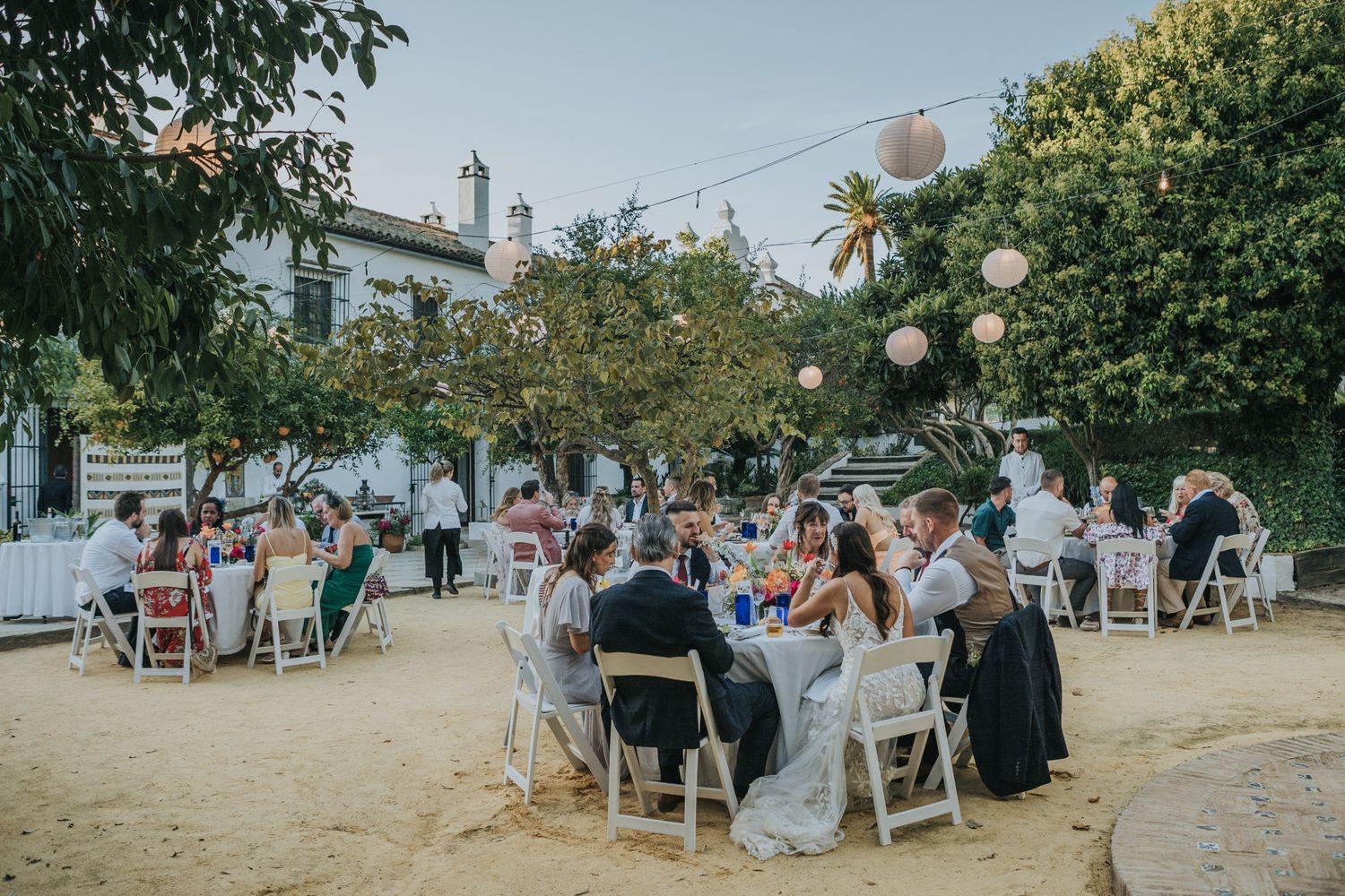 Outdoor garden reception dining setup with white folding chairs and tables under string lights and green foliage.